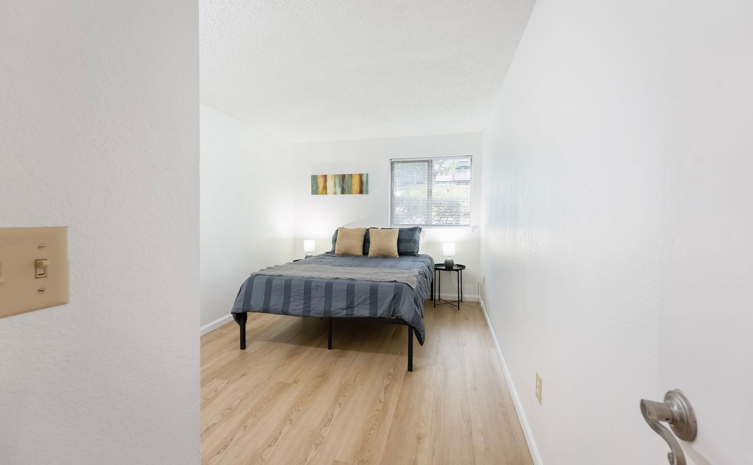 Minimalist bedroom with a queen bed, light hardwood floors, white walls, and a bright window providing natural light.