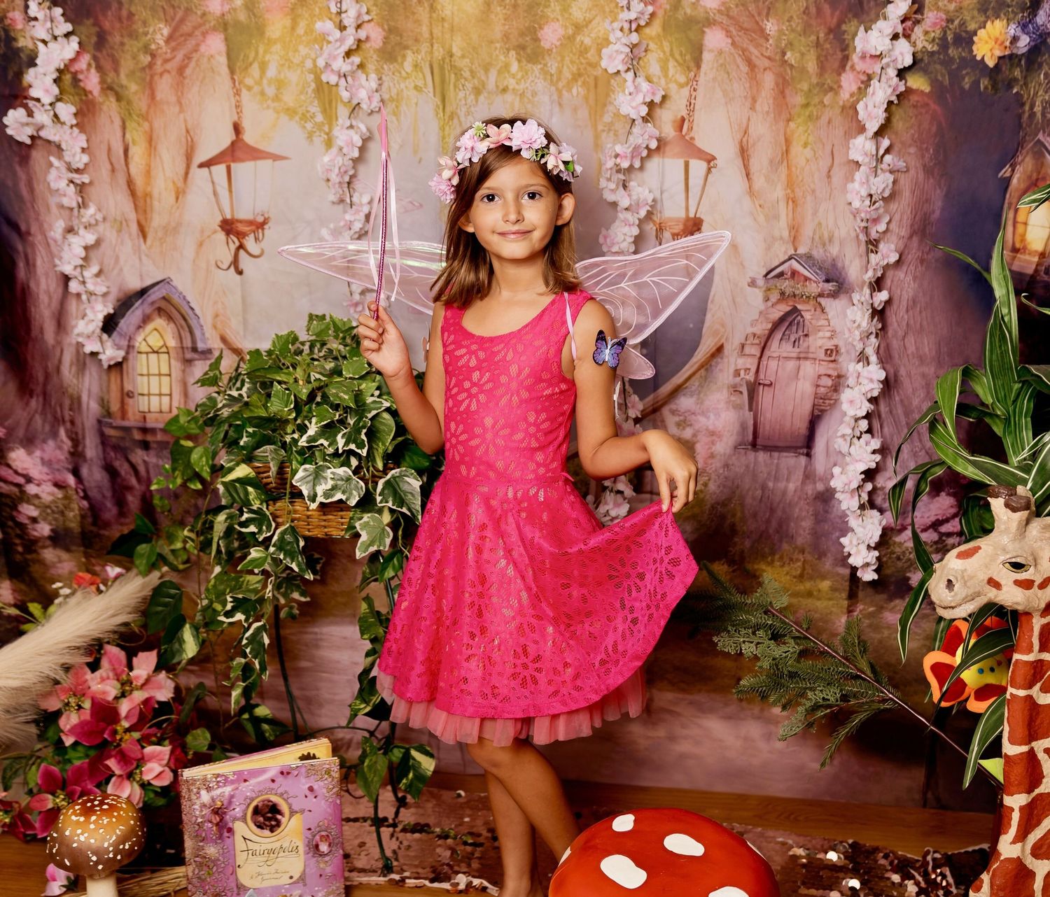 Young girl in pink fairy dress with wings and flower crown poses in magical enchanted garden photo backdrop.
