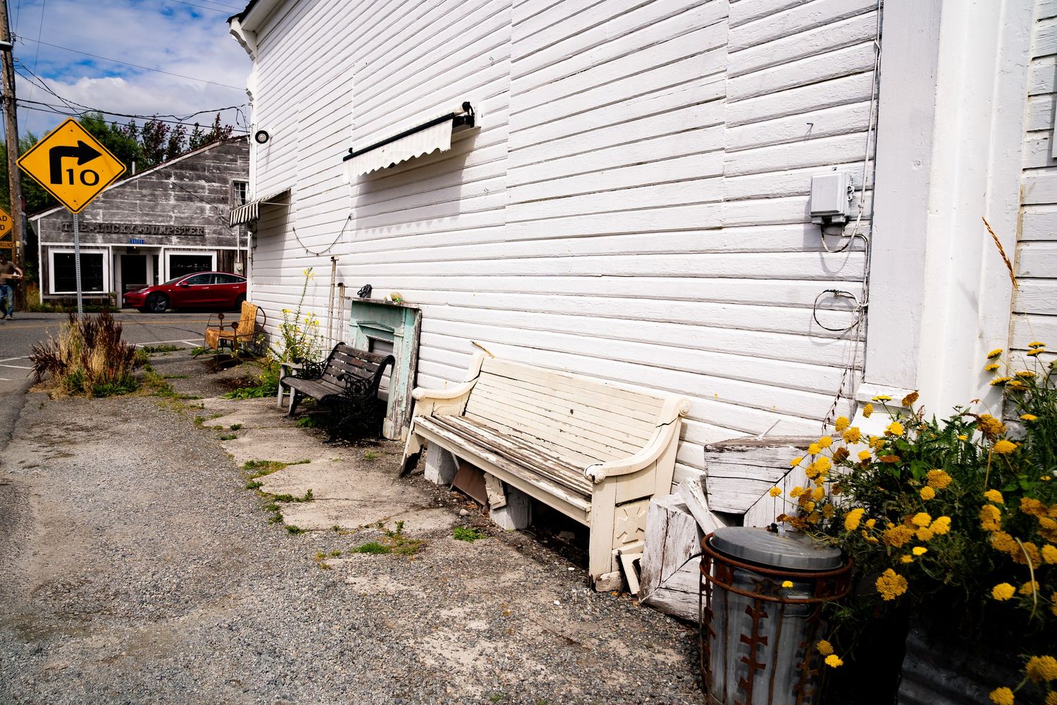White benches and yellow flowers line the side of a white wooden building beside a gravel lot.