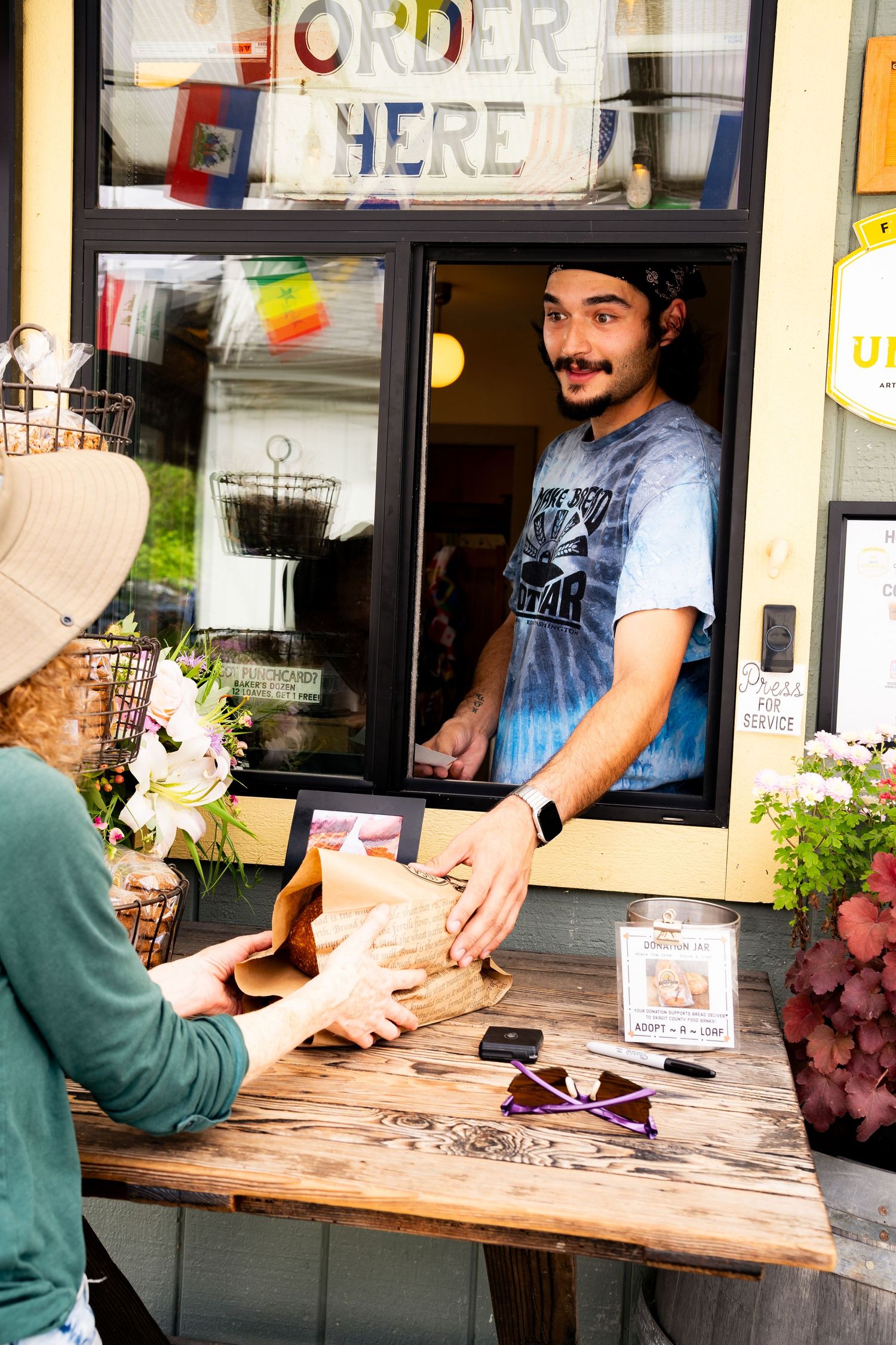 Young man serving a customer through a food stall window with an Order Here sign at an outdoor cafe.