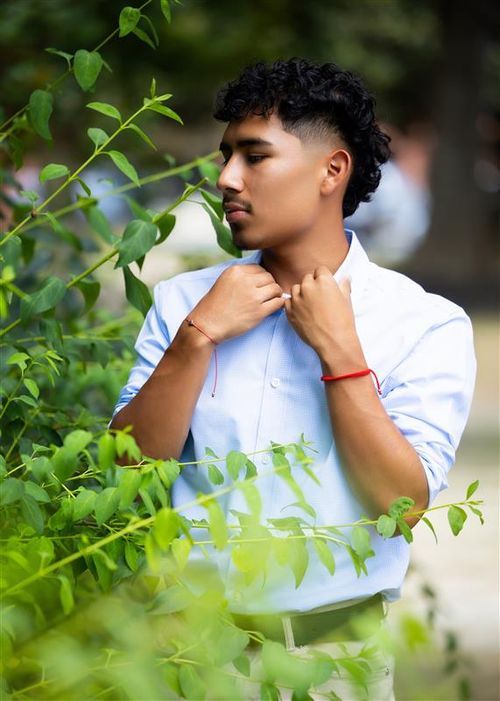 Young man in light blue shirt adjusting collar outdoors, surrounded by lush green foliage in natural setting.
