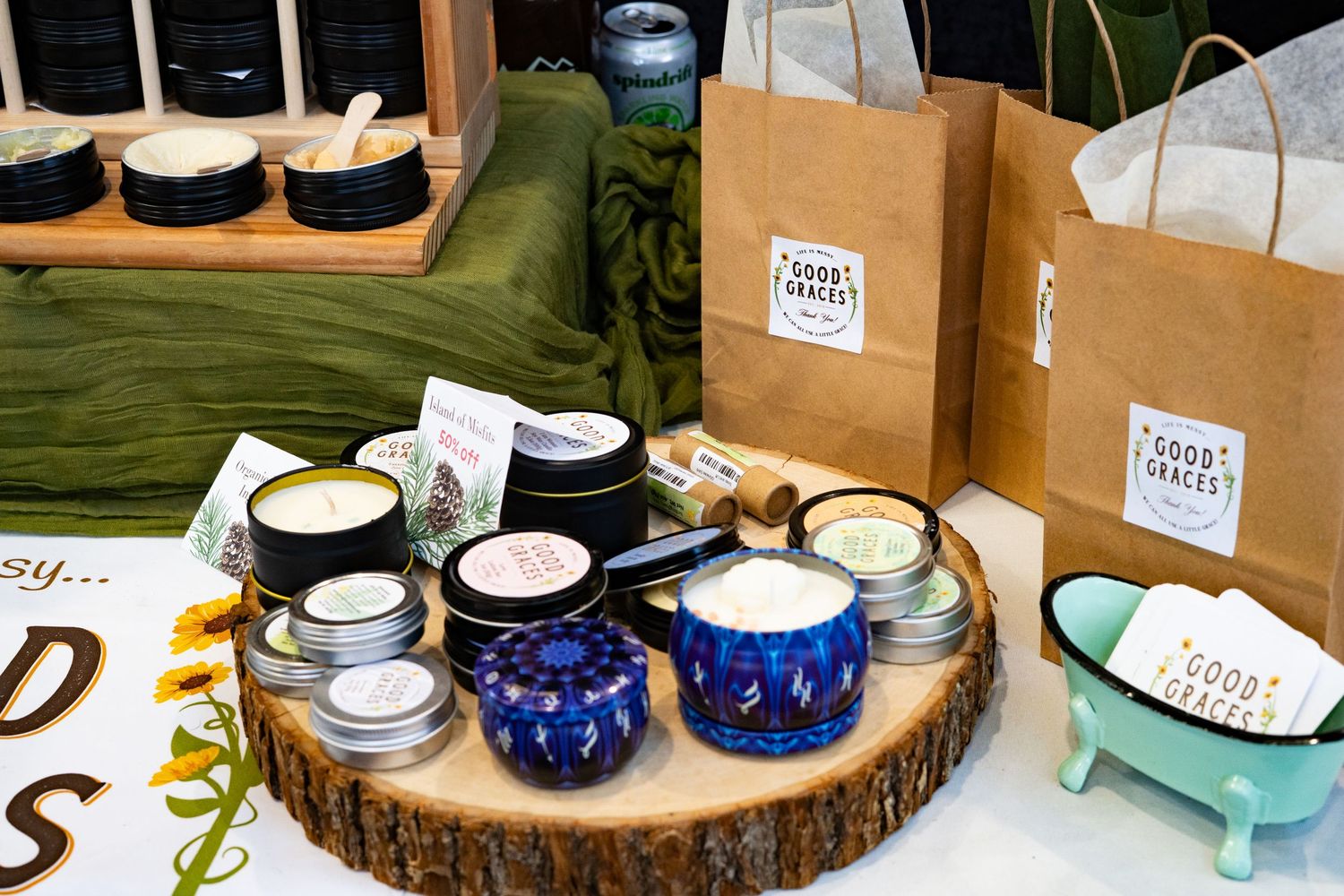 Artisan candles and bath products displayed on a wood slice with kraft paper gift bags at a craft fair.