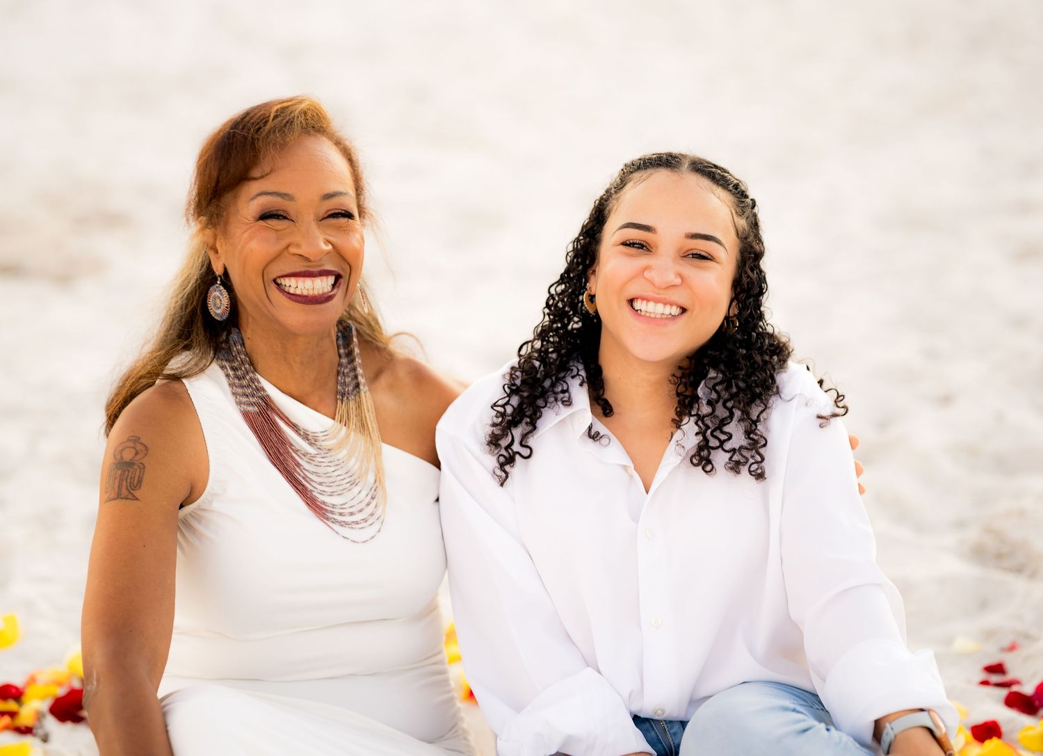 Two people sitting together and smiling on a beach in white outfits during a sunny day.