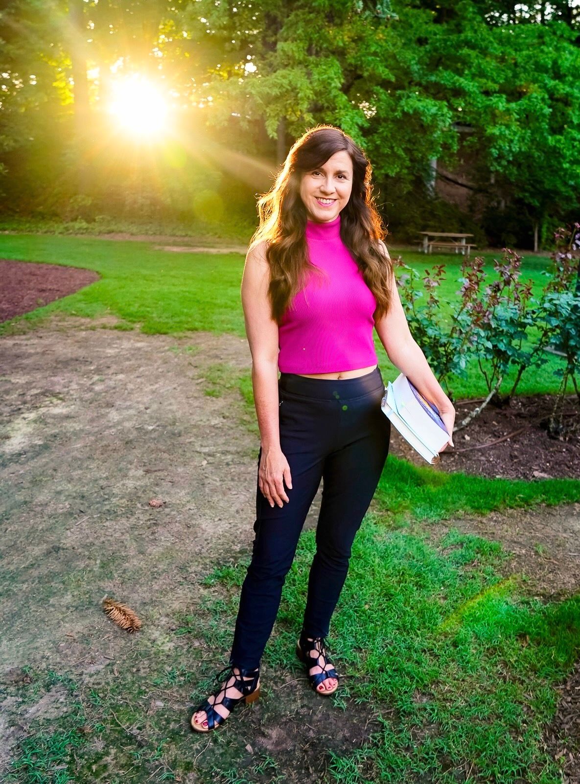 A person in a pink sleeveless top and dark pants poses outdoors in a sunlit park setting with green foliage.