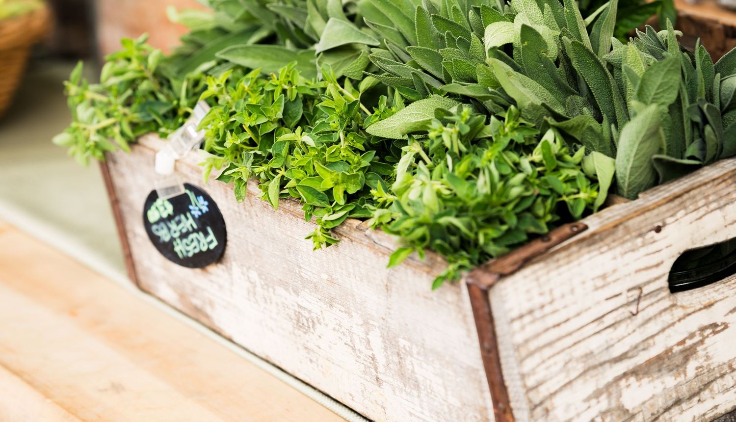 Fresh green herbs displayed in a rustic wooden crate at a farmers market, featuring sage and oregano.