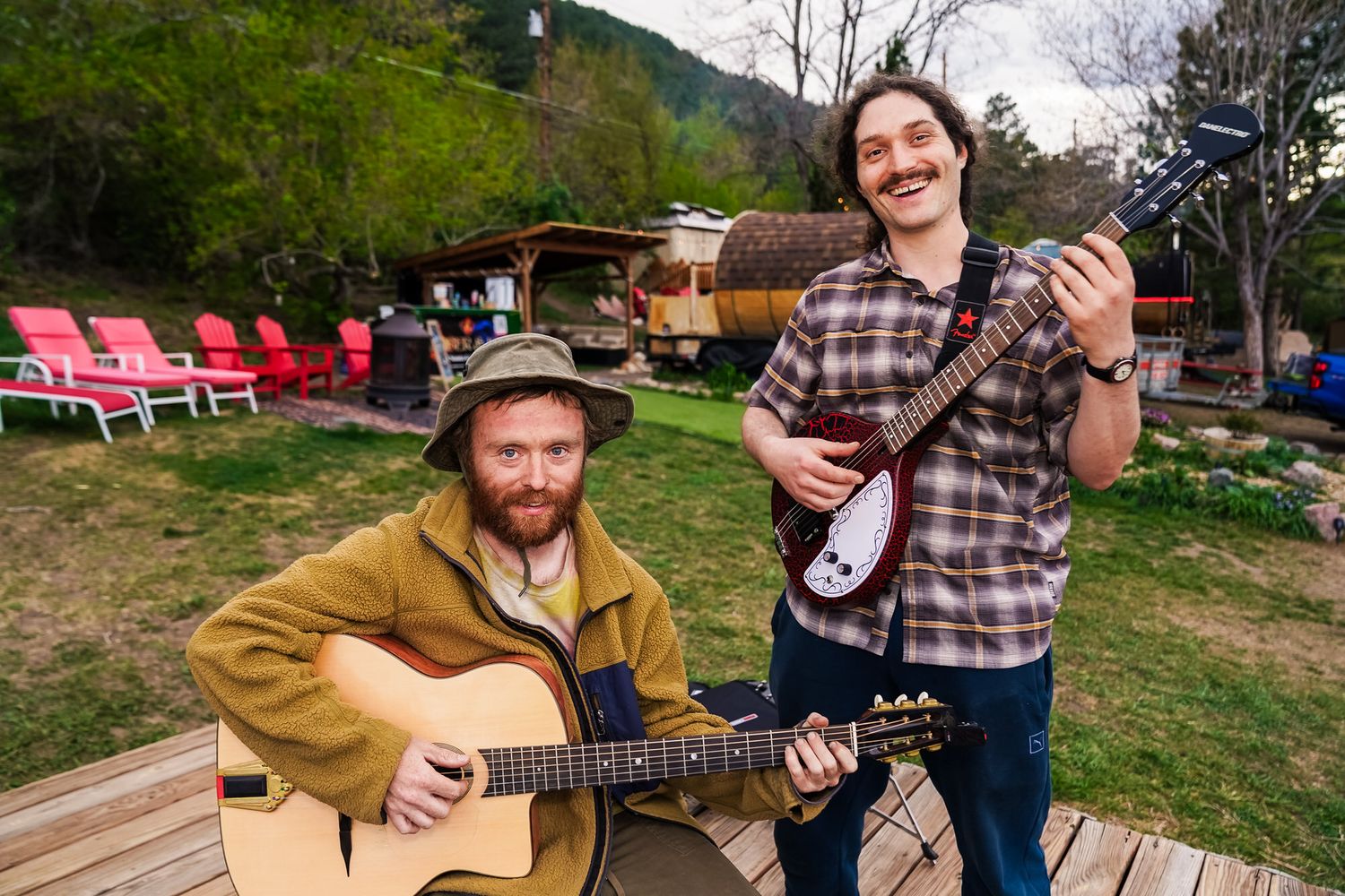 Two musicians smile outdoors, one playing acoustic guitar, one holding electric guitar, with colorful chairs and hills behind.