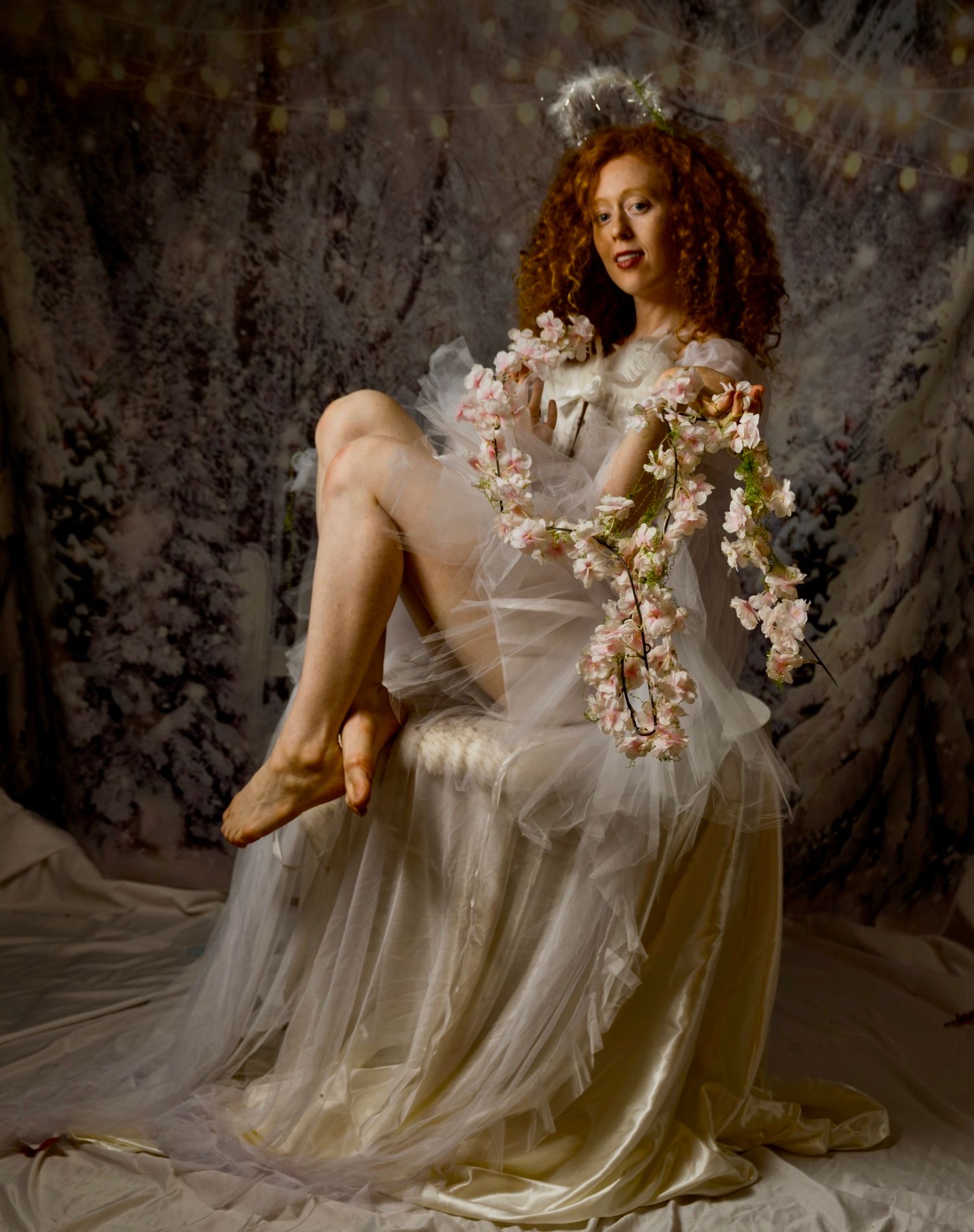 Woman in white tulle gown with curly red hair holding pink floral garland, wearing floral crown in artistic portrait.