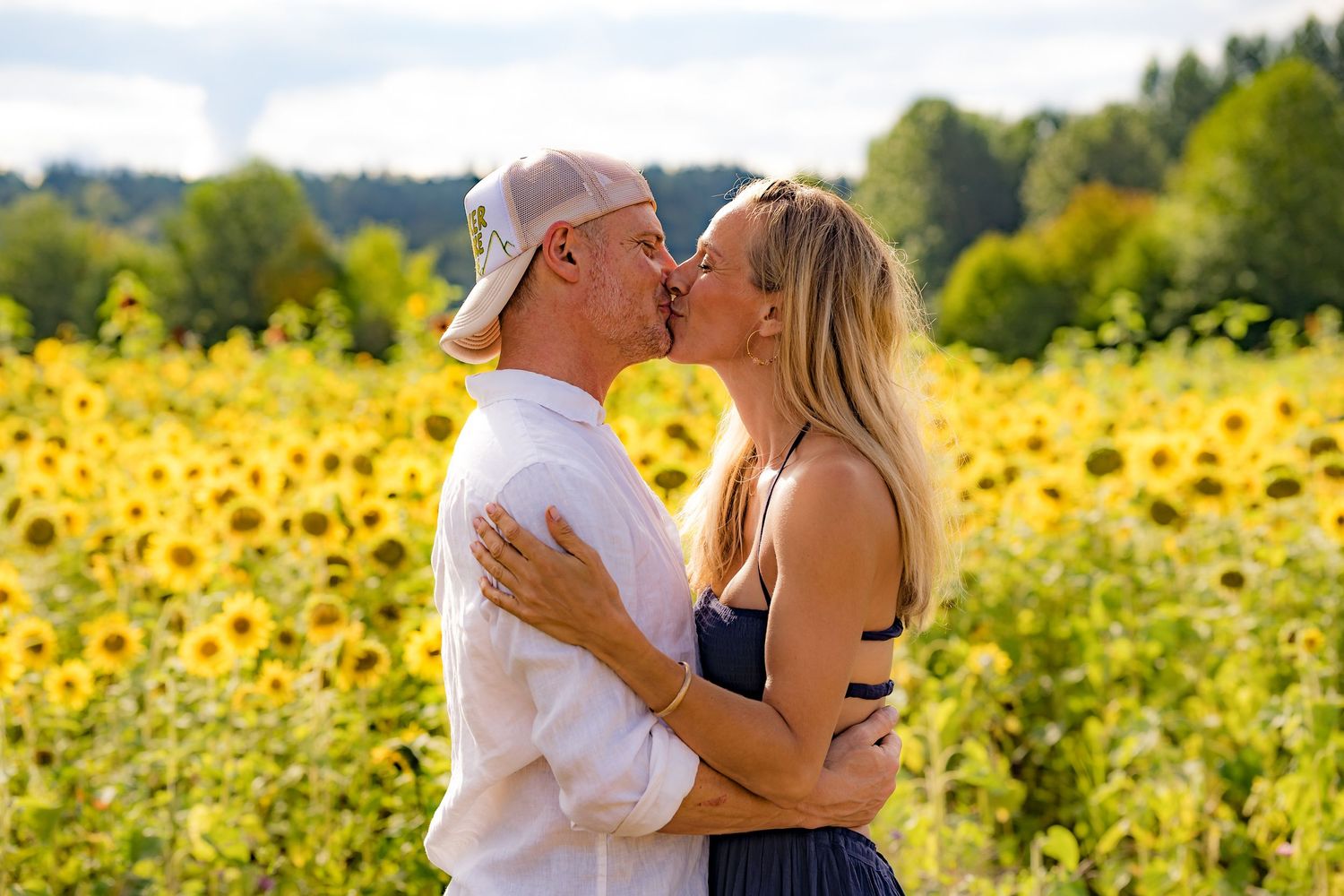 Couple kissing in a sunflower field on a sunny day, surrounded by bright yellow blooms and green trees.