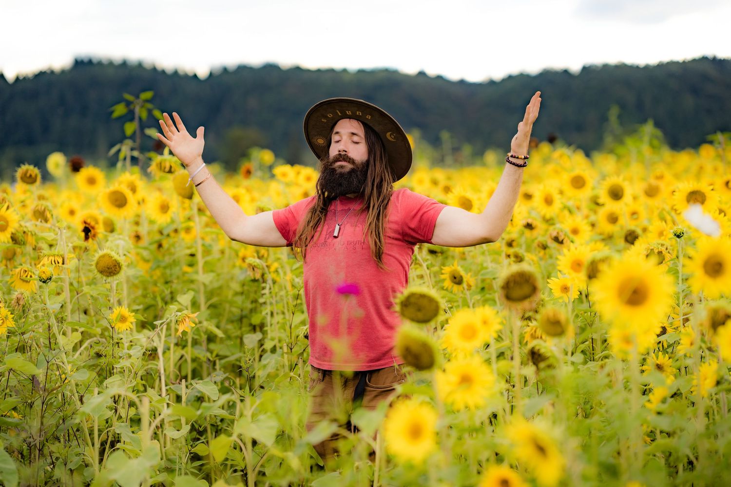 Bearded man with dreadlocks raises arms joyfully in a vibrant sunflower field wearing a red shirt and brown hat.