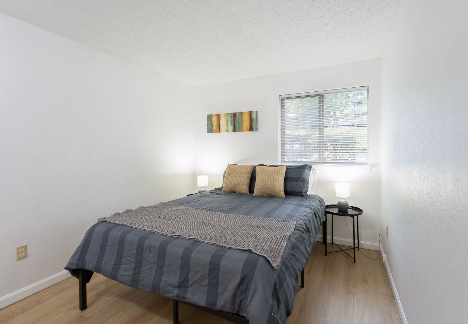 Bright bedroom with queen bed, gray striped bedding, wood floors, white walls, and a window with natural light.