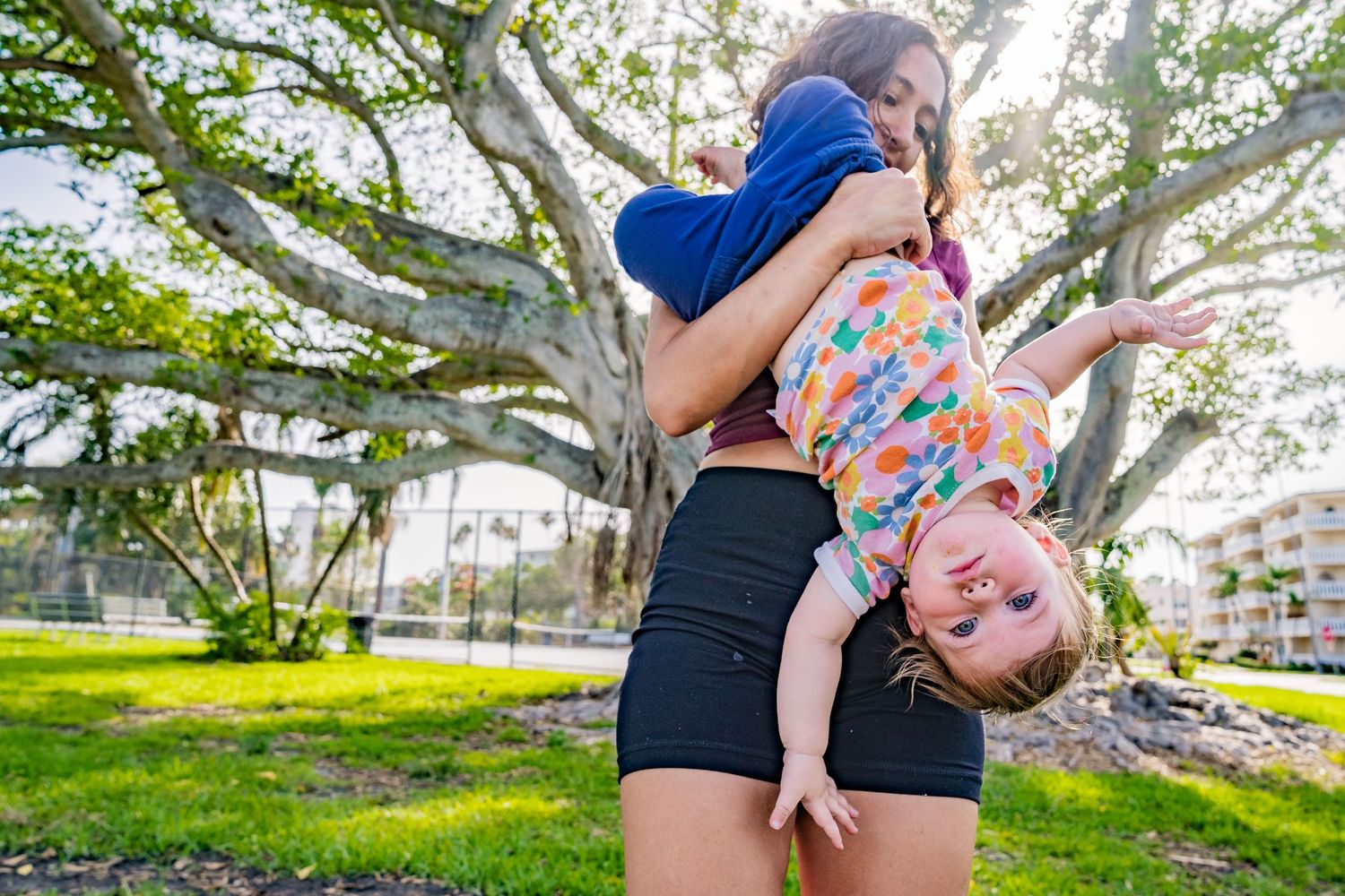 A photo of a mom hanging baby girl upset down with trees and grass behind them.