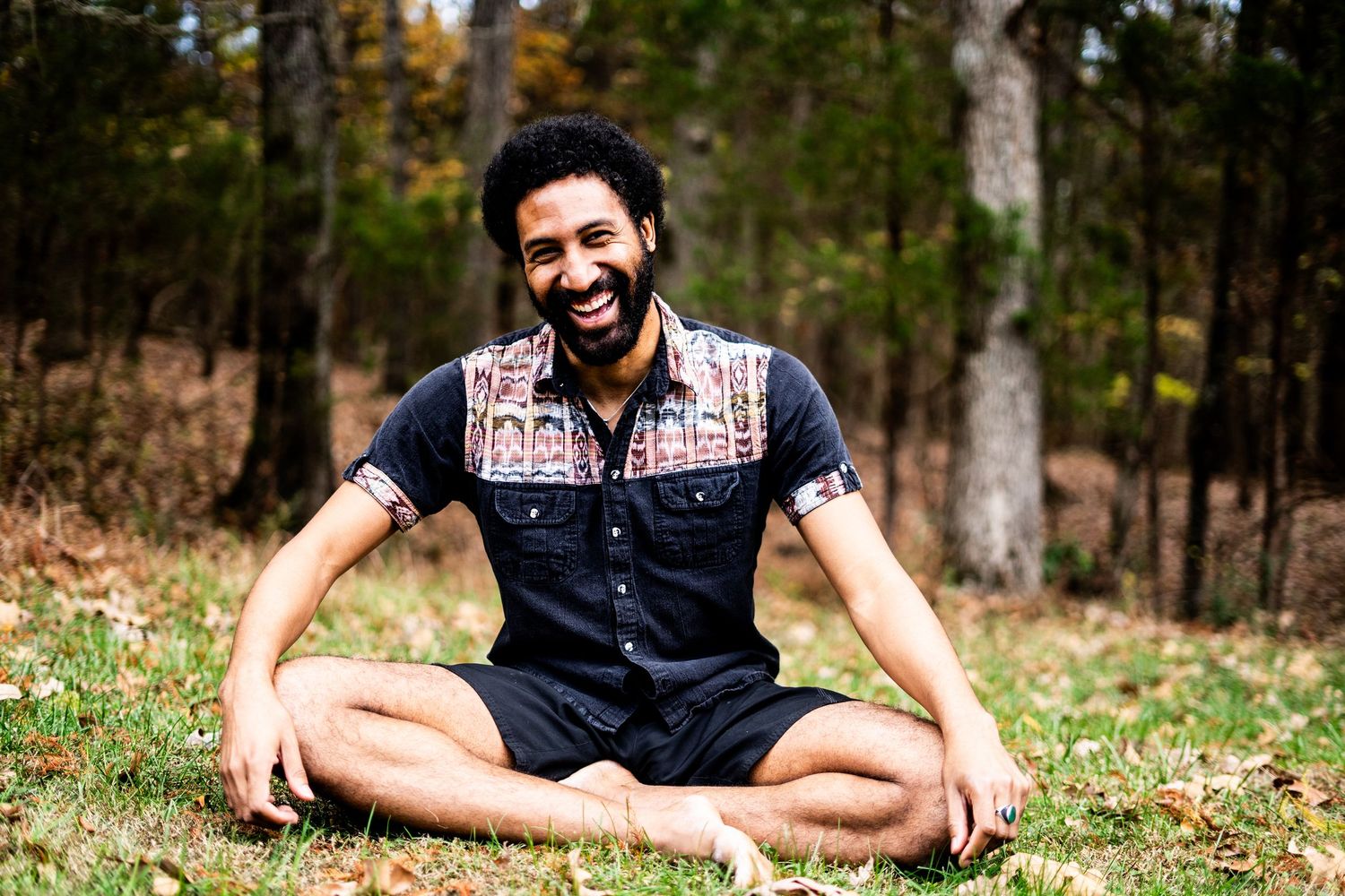 Smiling man sitting cross-legged on grass in a forest, wearing a patterned short-sleeve shirt, laughing joyfully.