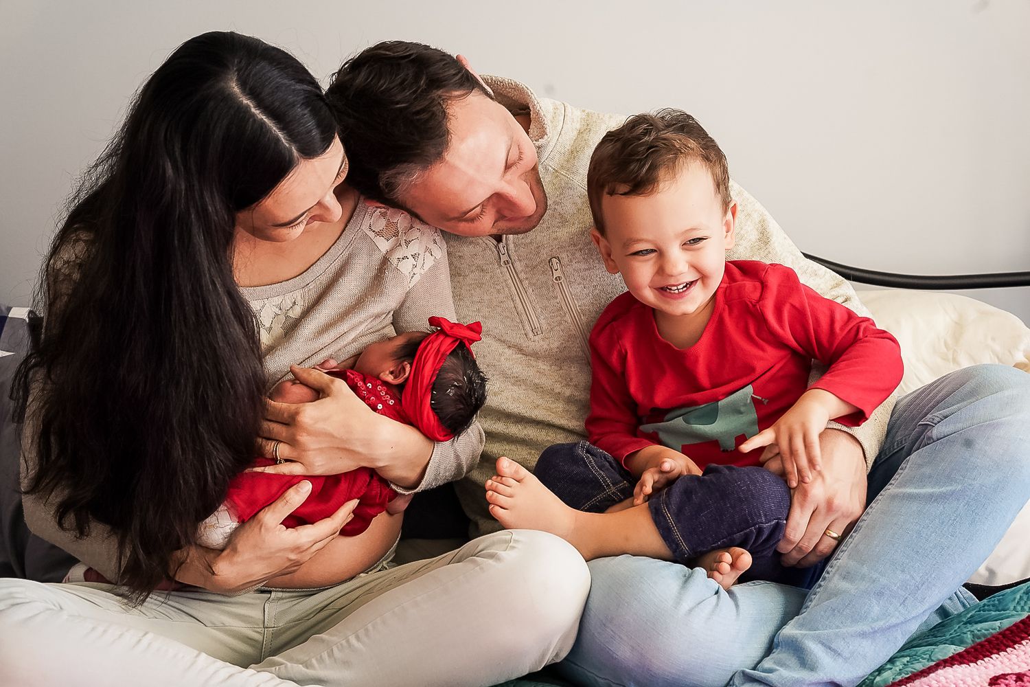 Happy family with parents and two young children, including a newborn baby, sitting together on a cozy bed smiling.