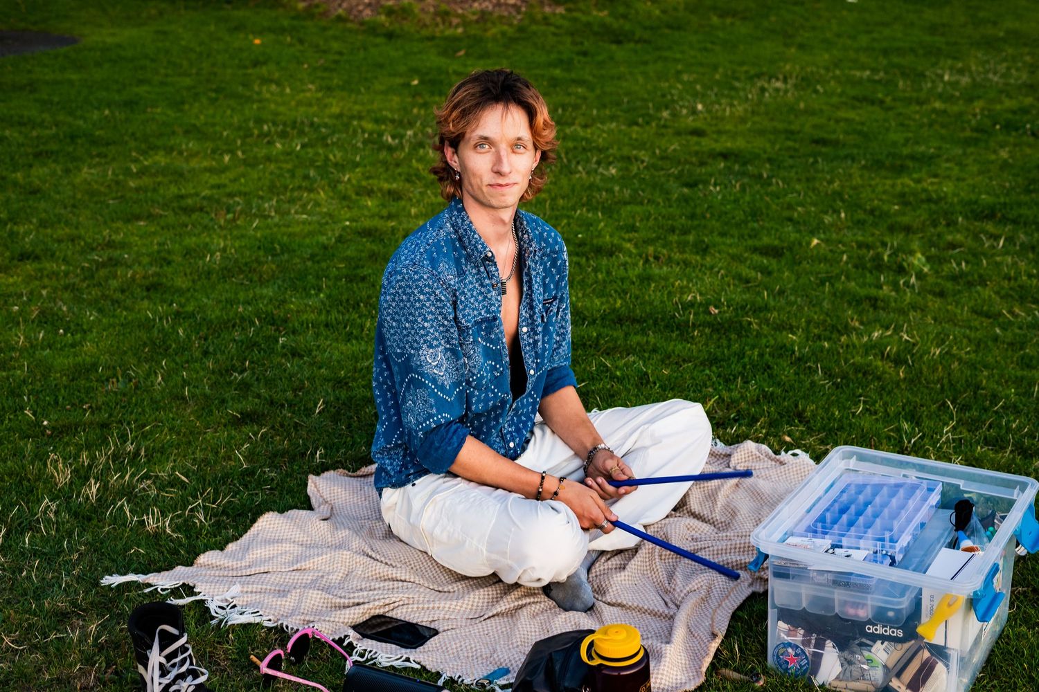 Young artist sitting cross-legged on blanket in green park, holding paintbrush beside art supply case.