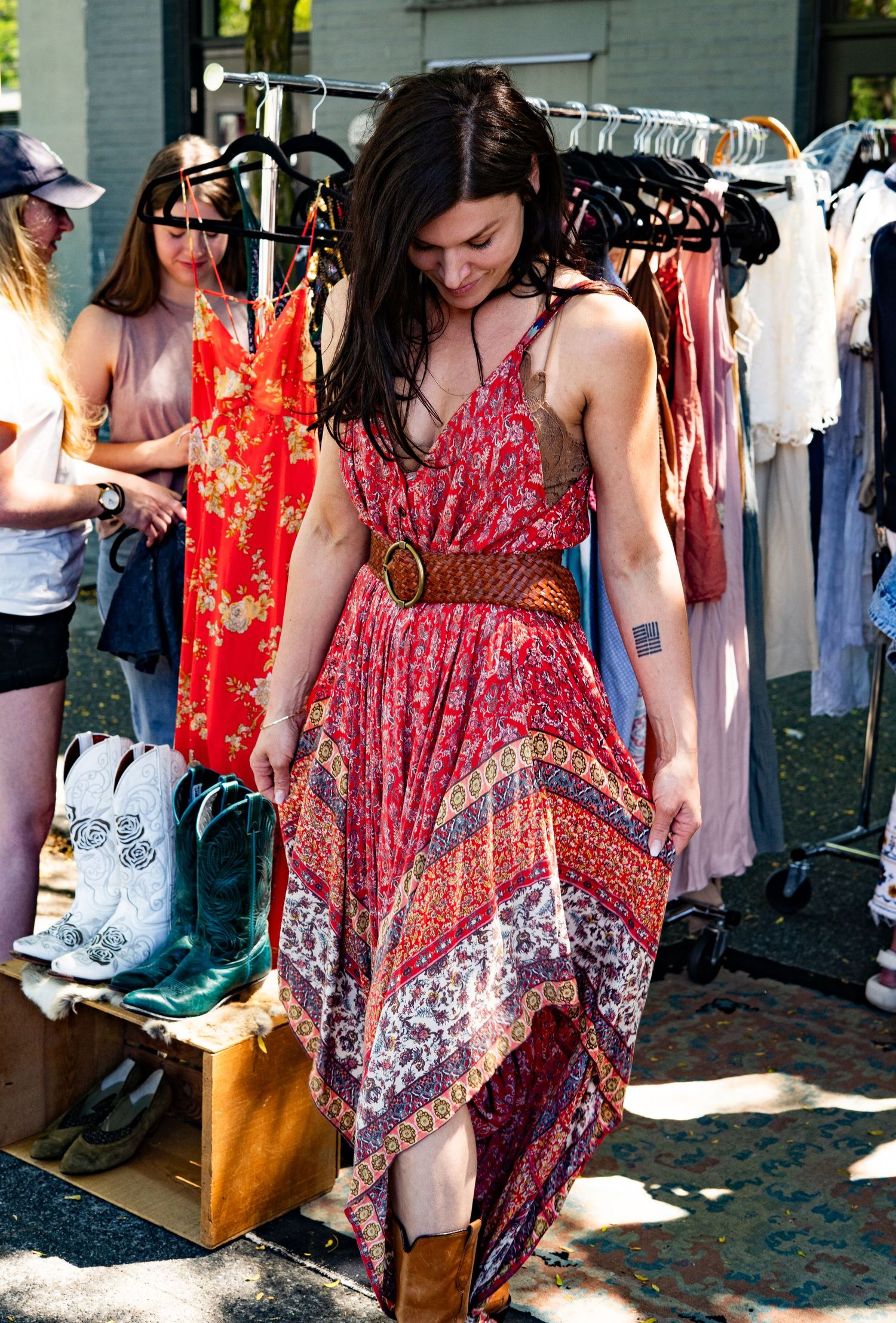 Woman in red boho maxi dress with brown belt and cowboy boots shopping at outdoor vintage clothing market.