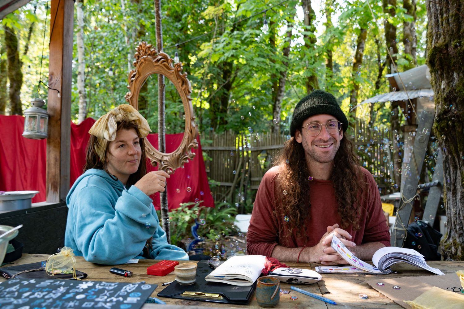 Two people sit outdoors at a table with books and a mirror, surrounded by lush green trees and red fabric.