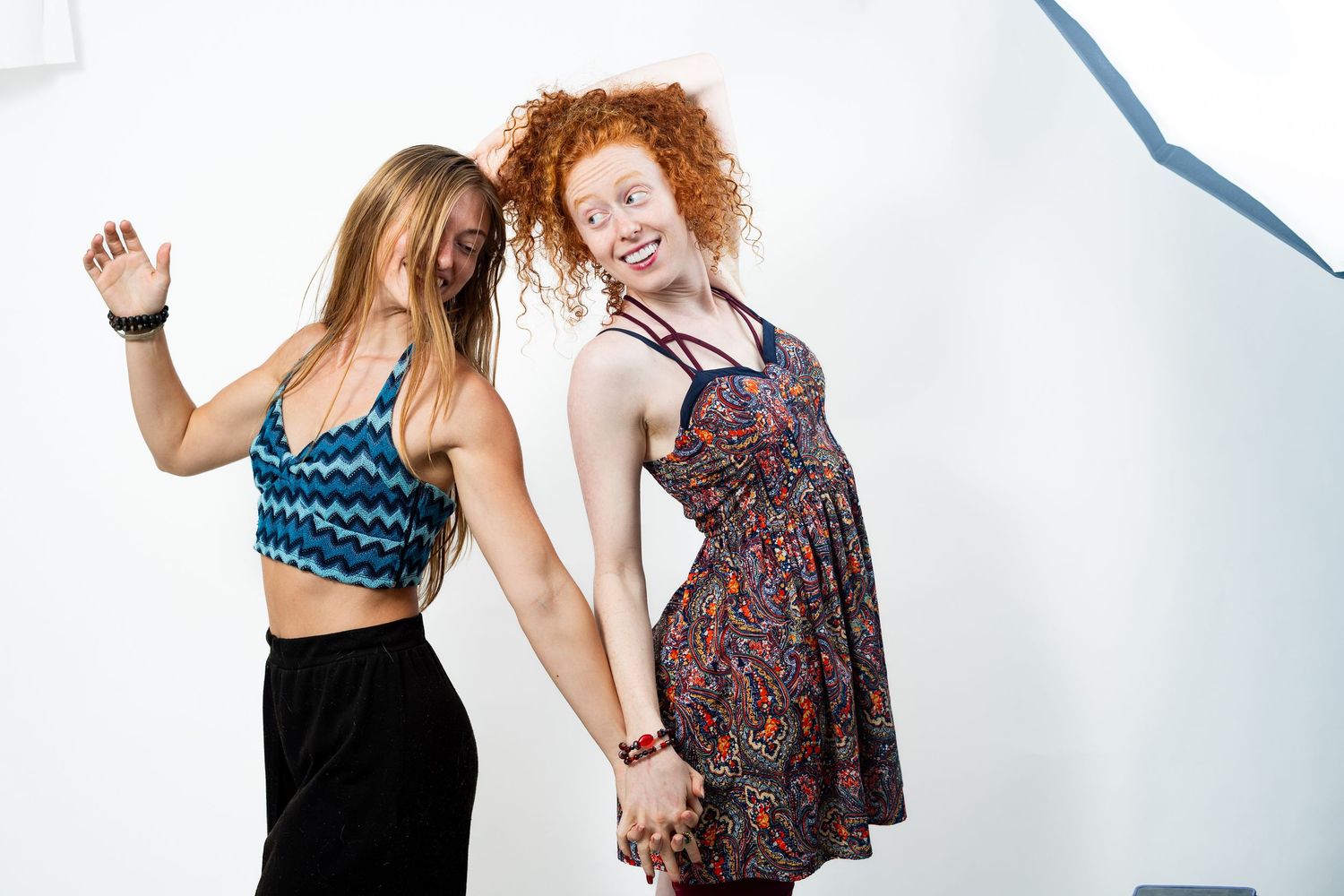 Two young women dancing and holding hands, one in a blue crop top and one in a floral dress, against a white background.