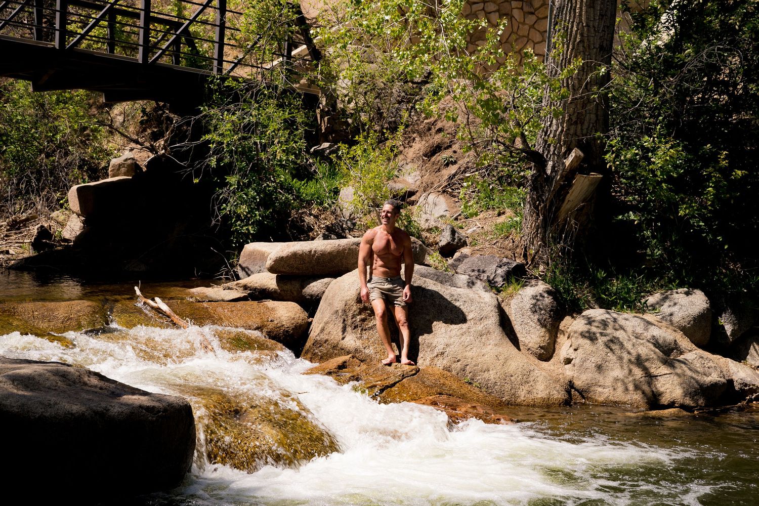Shirtless man standing on rocks beside a rushing mountain stream near a bridge.