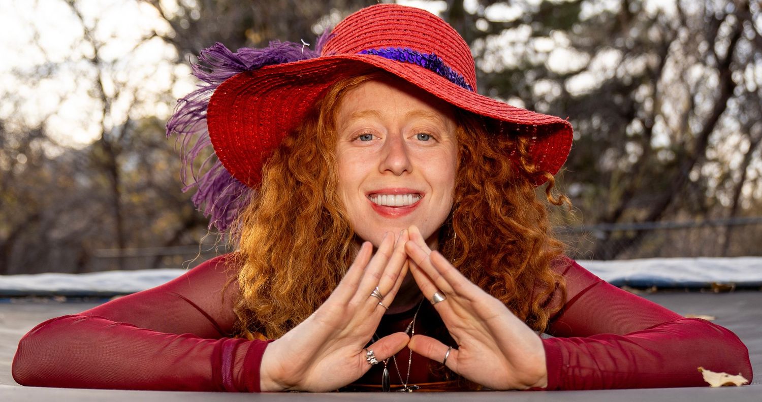 A person in a red hat lies outdoors on fallen autumn leaves while smiling at the camera.