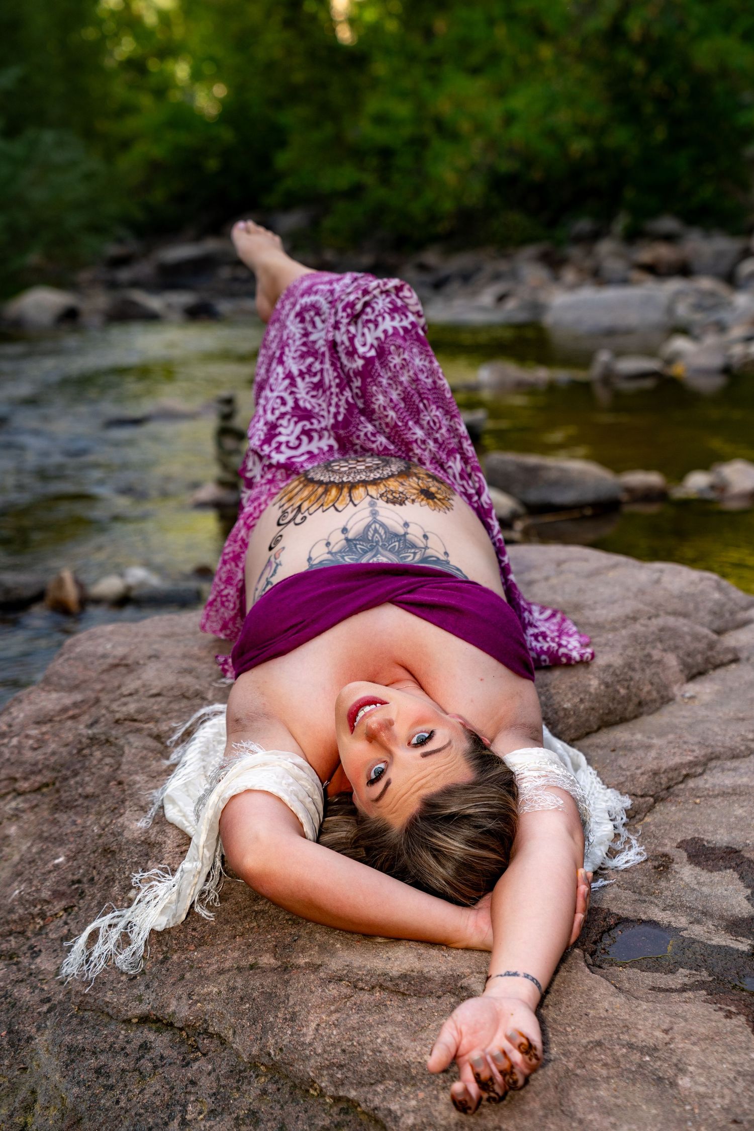 A person in a purple dress lies peacefully on a large rock by a stream in a natural outdoor setting.