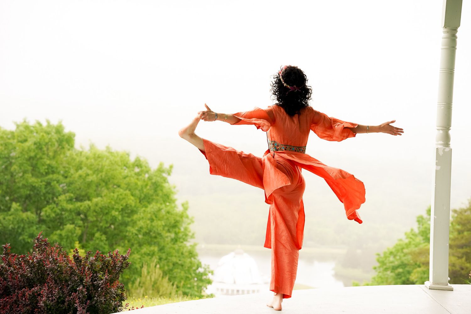 Woman in orange outfit gracefully balancing on one leg outdoors with trees in background.