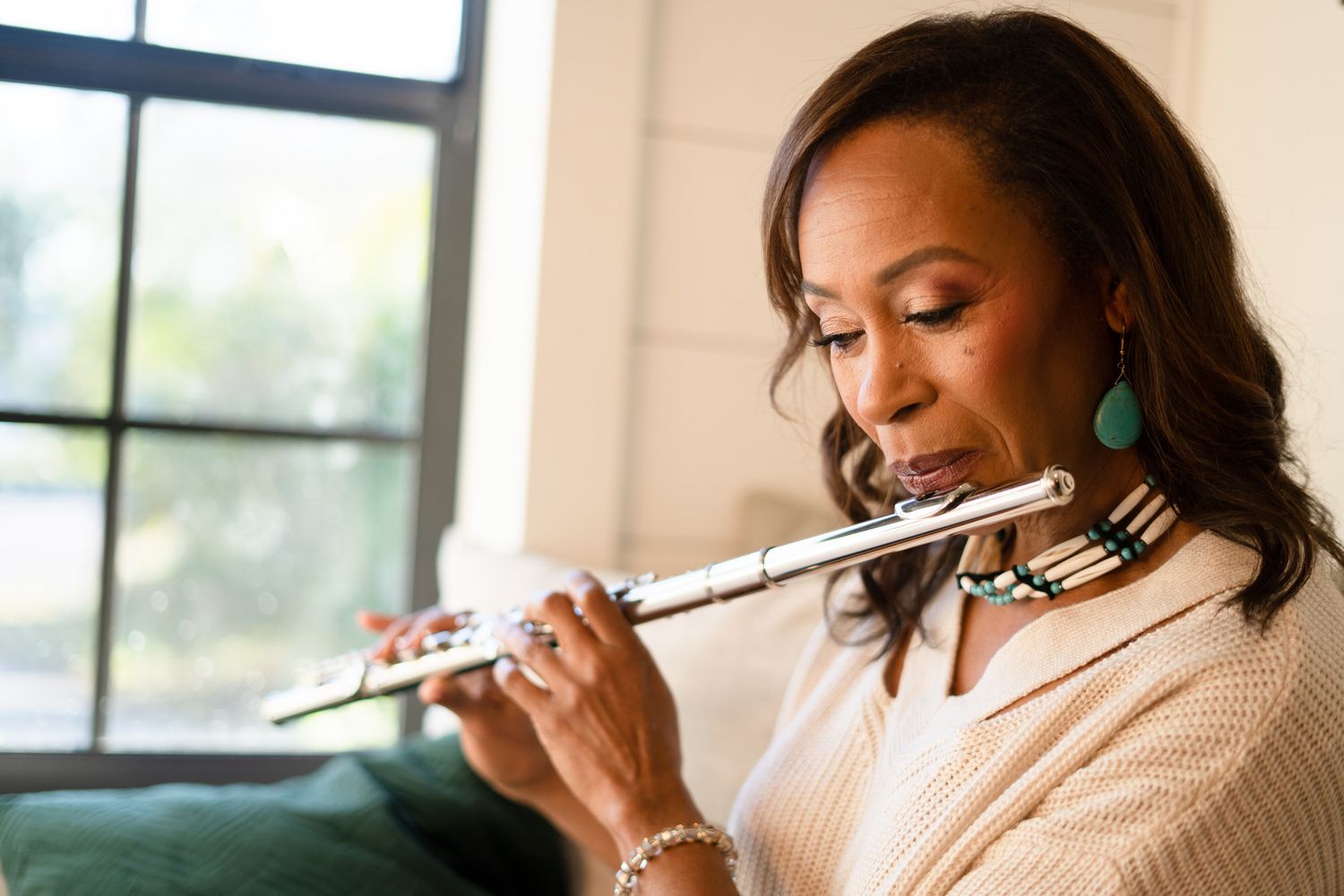 Woman playing a silver flute wearing teal earrings and a beige sweater in a bright indoor setting.