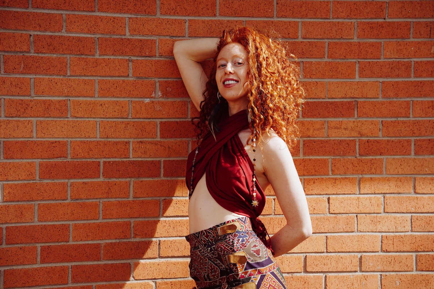 Young woman with curly red hair poses confidently against a brick wall in a crop top and patterned pants.