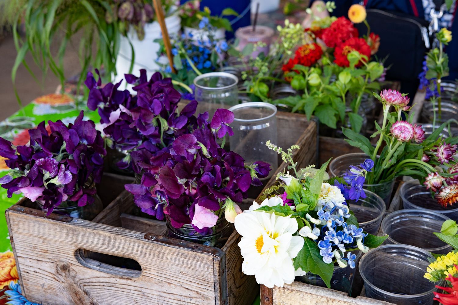 Colorful fresh flowers displayed in rustic wooden crates and glass jars at an outdoor flower market stall.