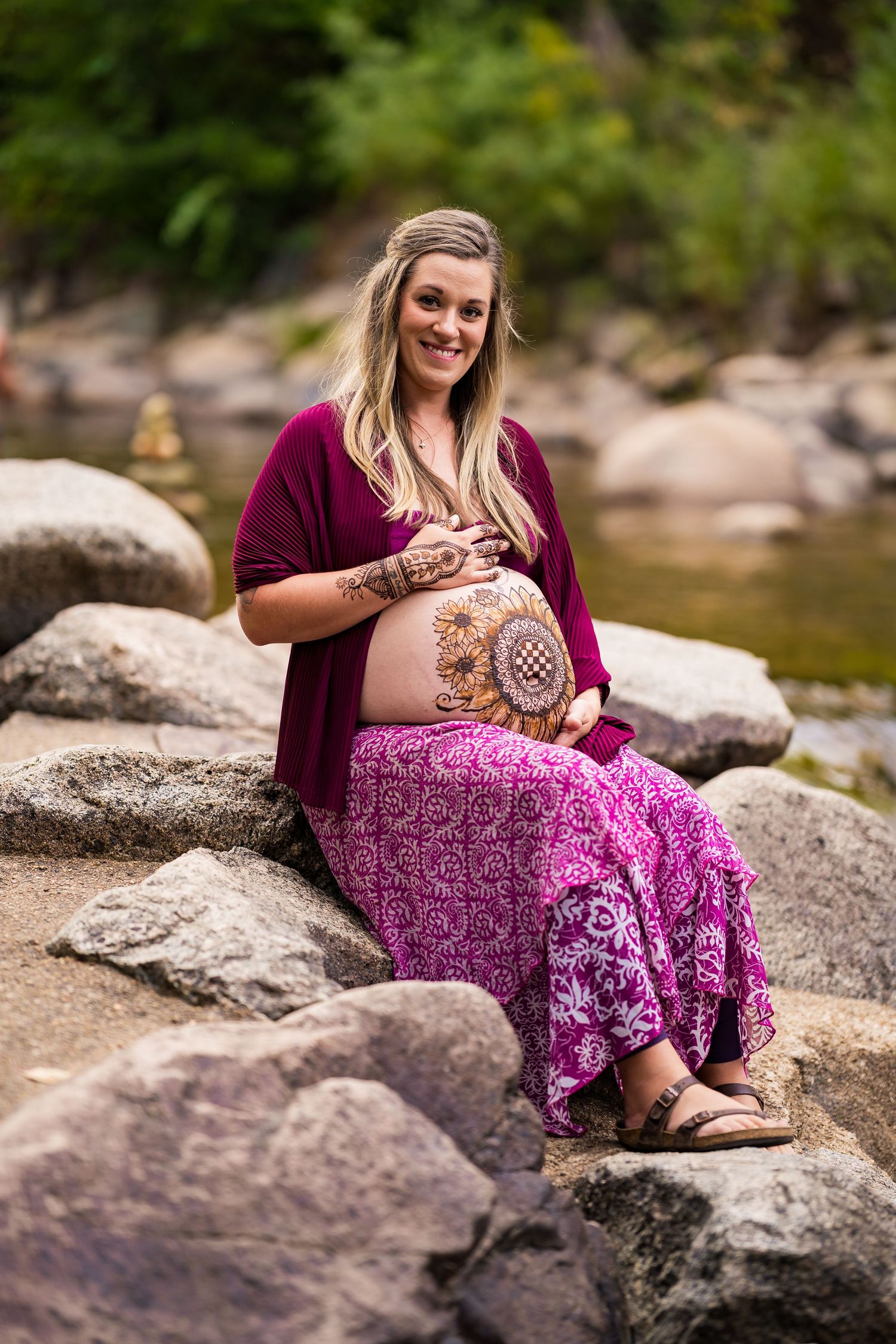 A woman in a purple dress sits on rocks by a river, showcasing a maternity photoshoot in nature.