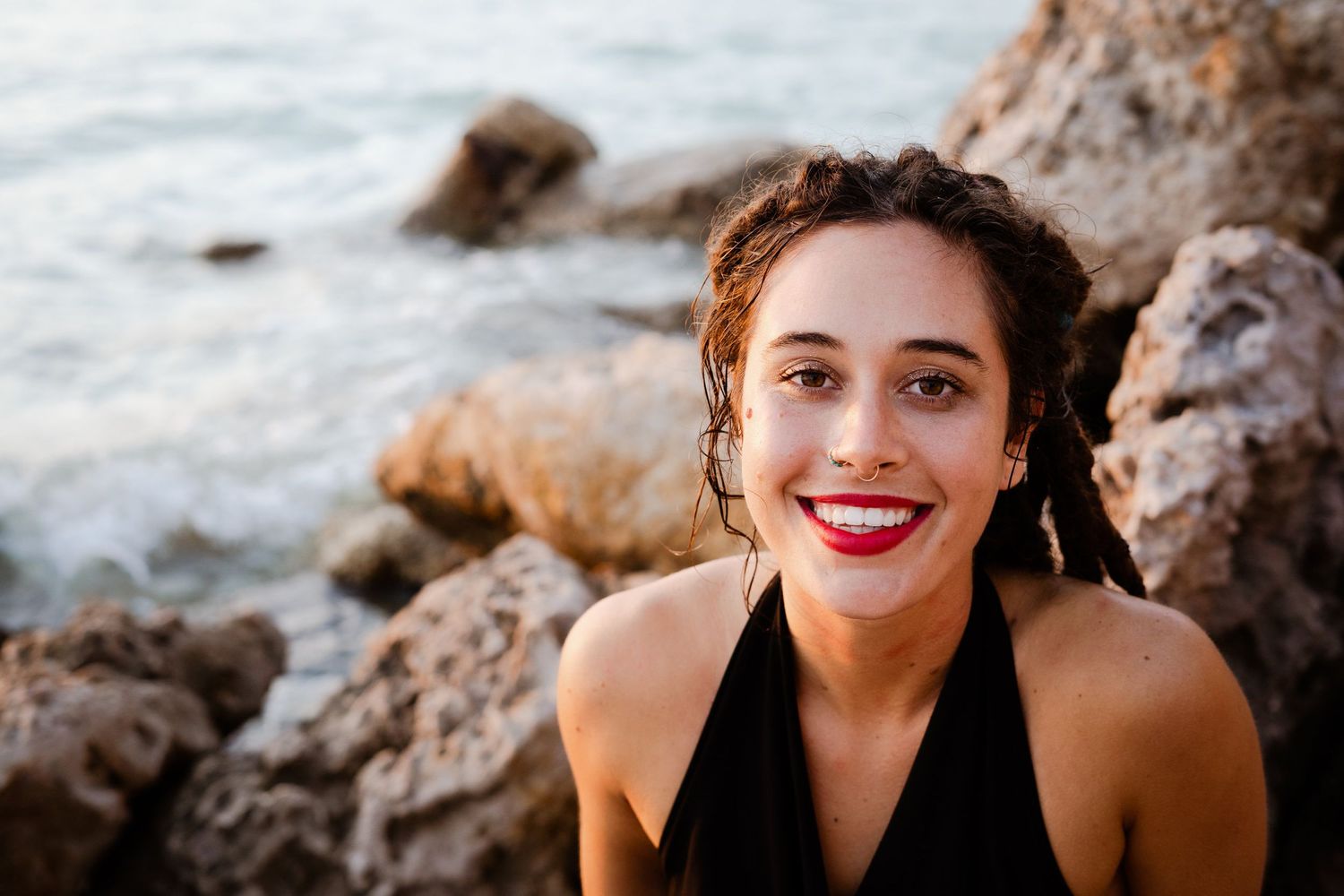 A girl with brown hair smiling at the beach, wearing red lipstick.