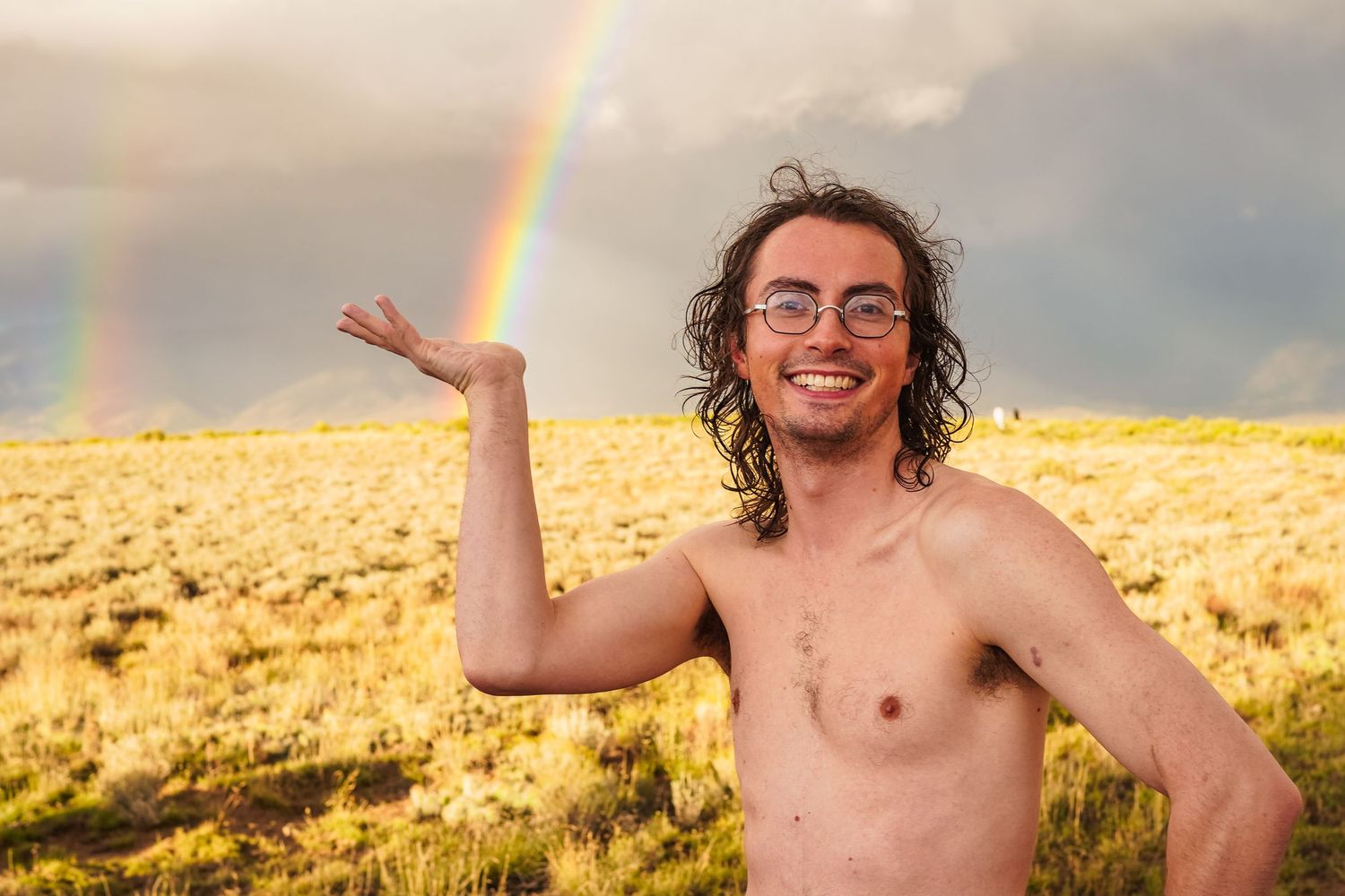 A photo of a shirtless guy with curly brown hair, he is holding a rainbow and smiling.