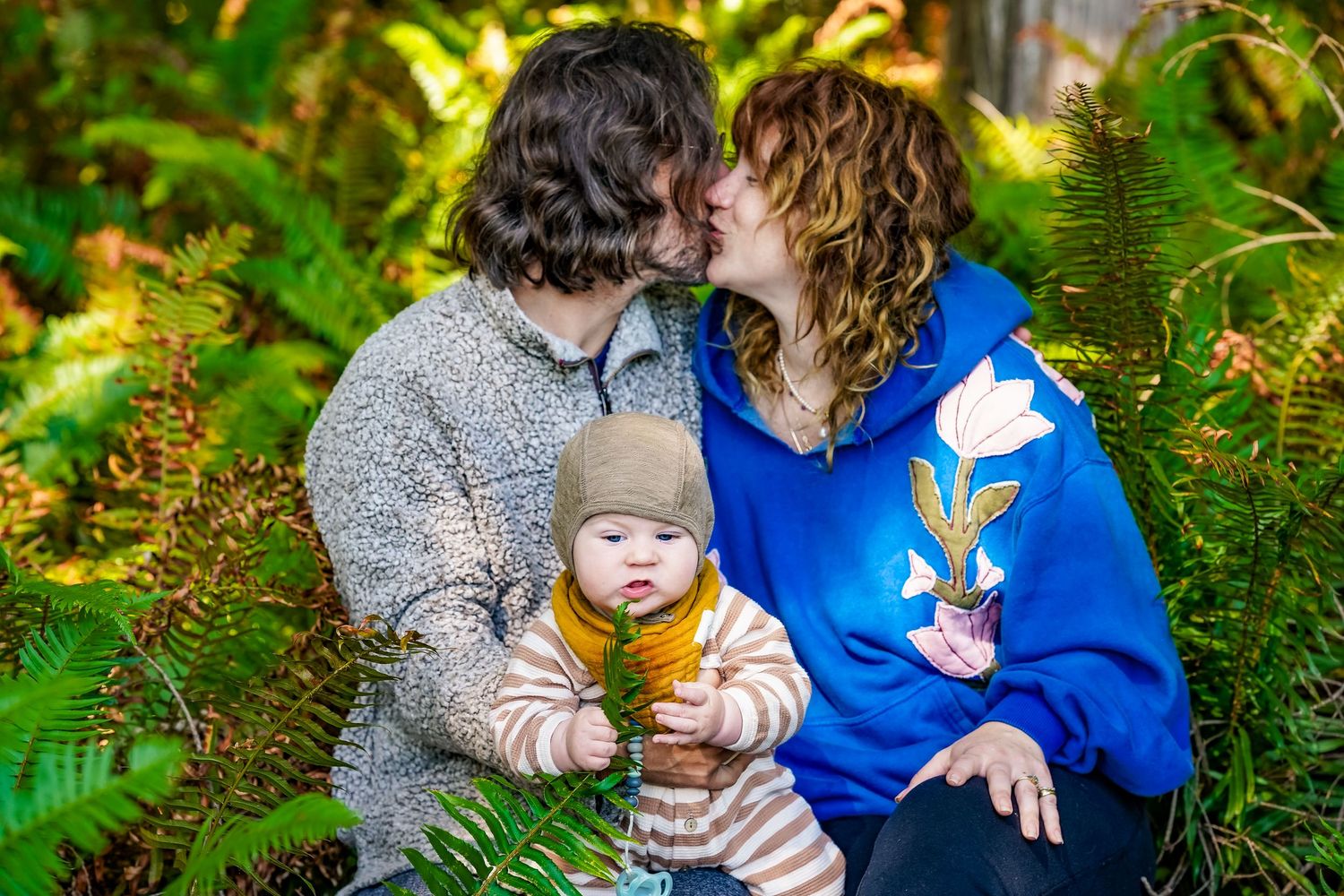 A couple kisses while holding their baby outdoors among lush green ferns in a forest setting.