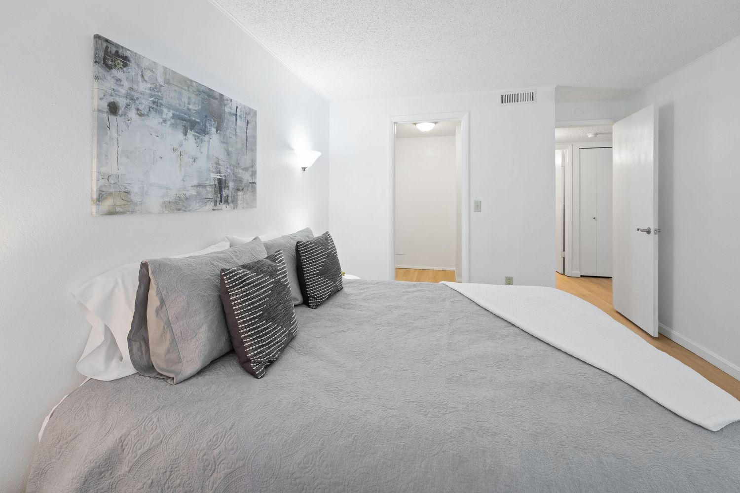 Modern bedroom with gray bedding, decorative pillows, abstract wall art, and white walls with hardwood flooring visible.