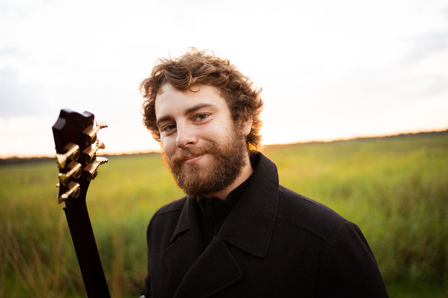 Bearded musician with curly hair holding a guitar outdoors in a field at dusk, wearing a black jacket.