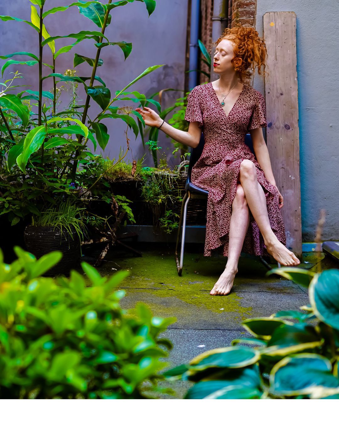 A redhead woman in a floral wrap dress sits barefoot in a lush garden courtyard, gazing thoughtfully to the side.