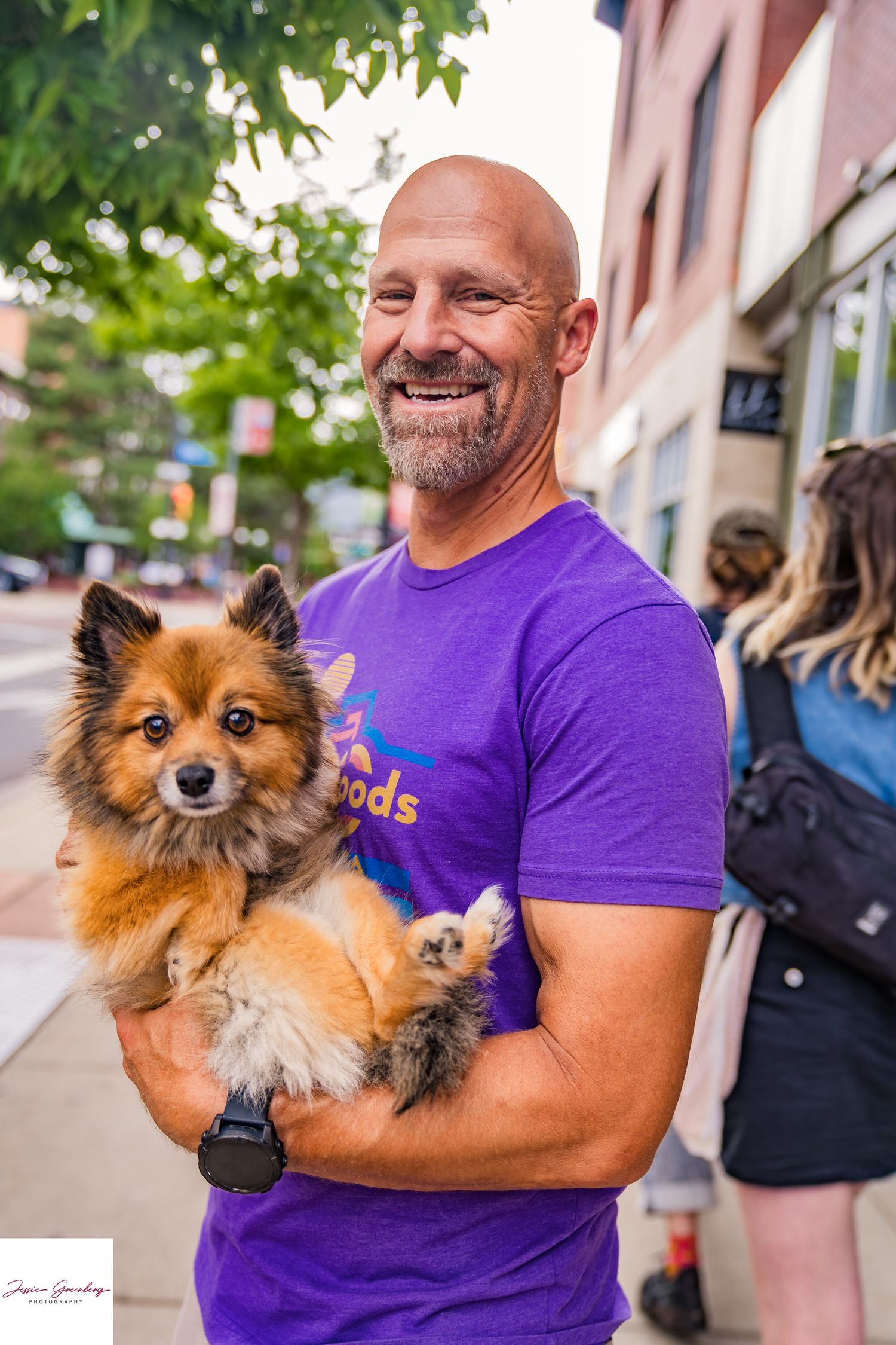 Bald smiling man in purple t-shirt holding a fluffy Pomeranian dog on city sidewalk.