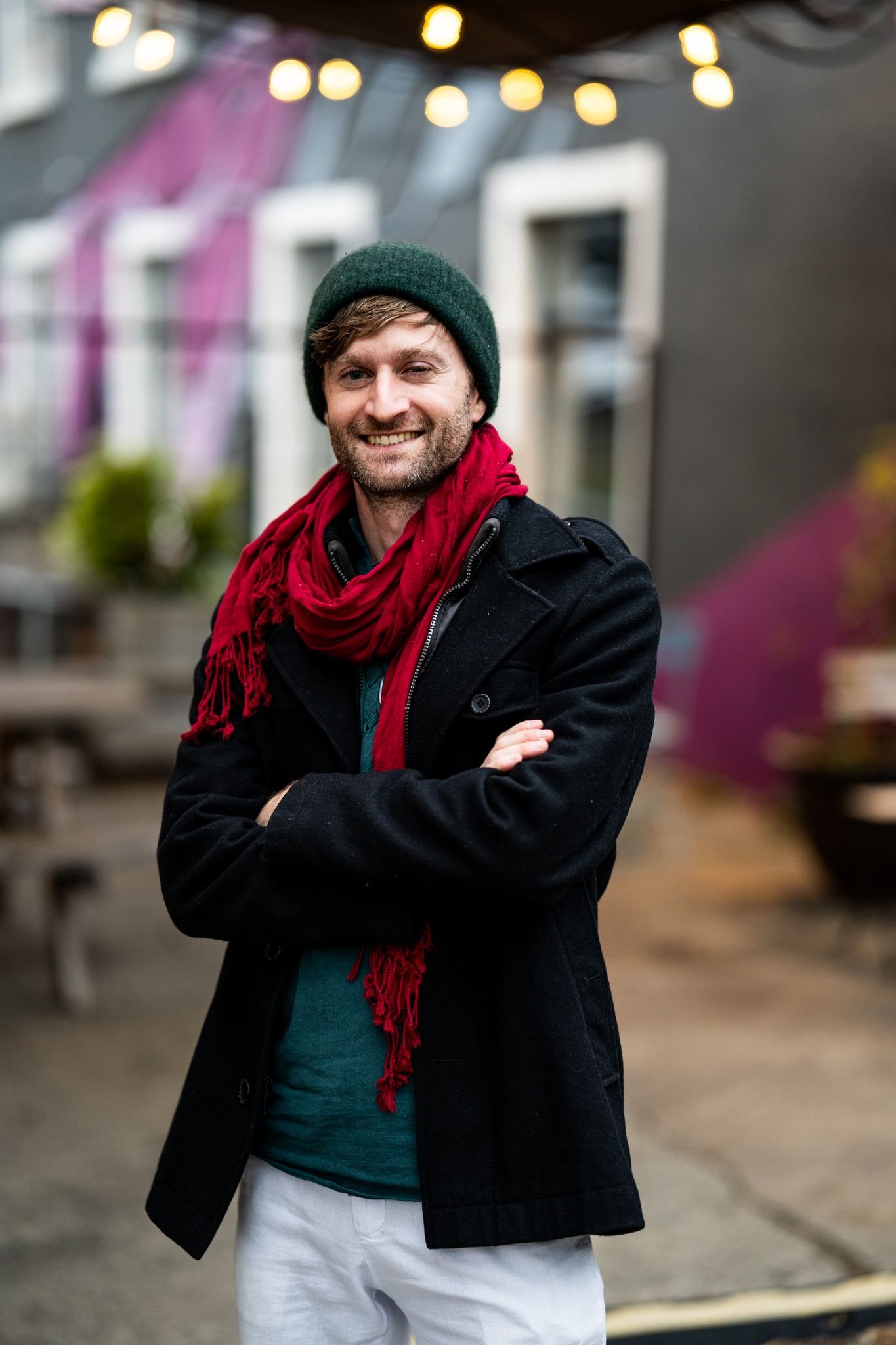 Smiling man with crossed arms wearing green beanie, red scarf, and black coat outdoors.