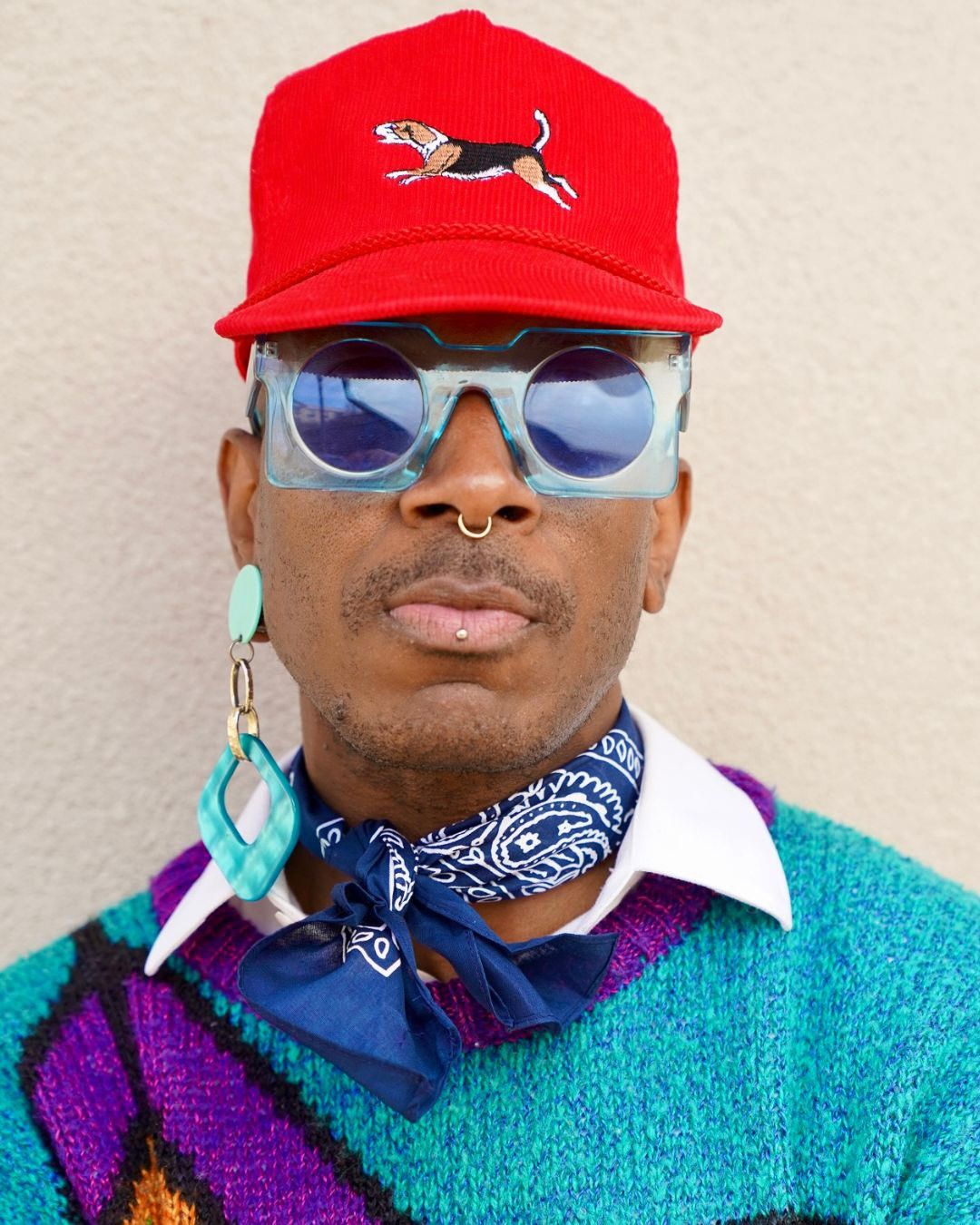Stylish person wearing red cap, blue sunglasses, nose ring, blue bandana, and colorful teal sweater against neutral background.