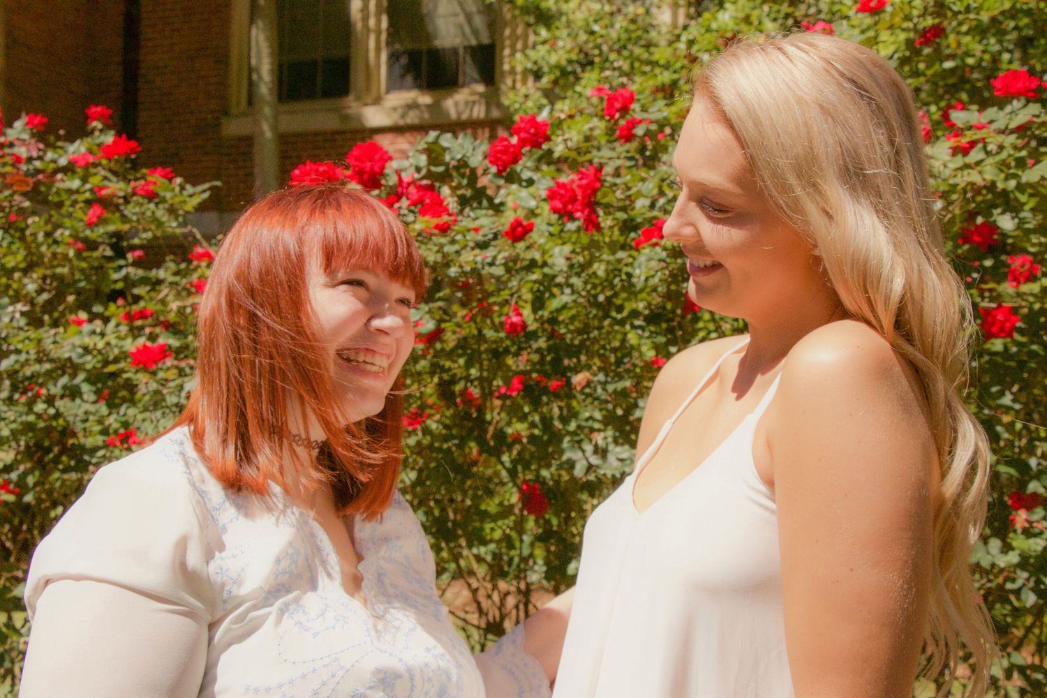 Two women with red and blonde hair smile at each other in front of blooming red roses outdoors.