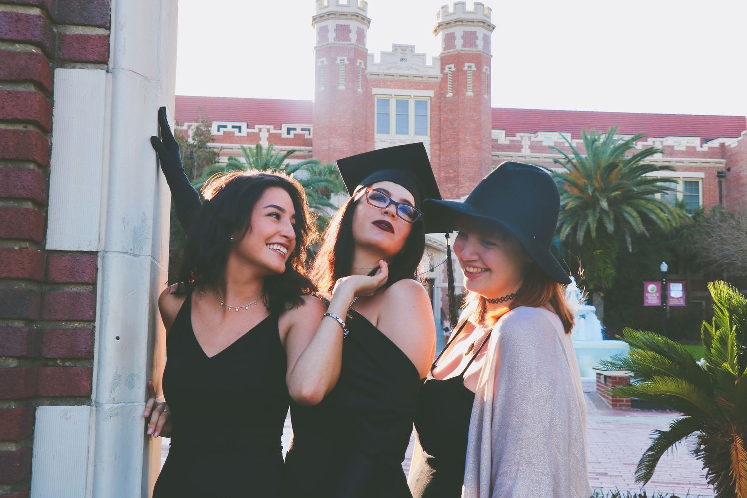 Three young women celebrating graduation outdoors, one wearing a cap and gown, smiling together near a brick building.