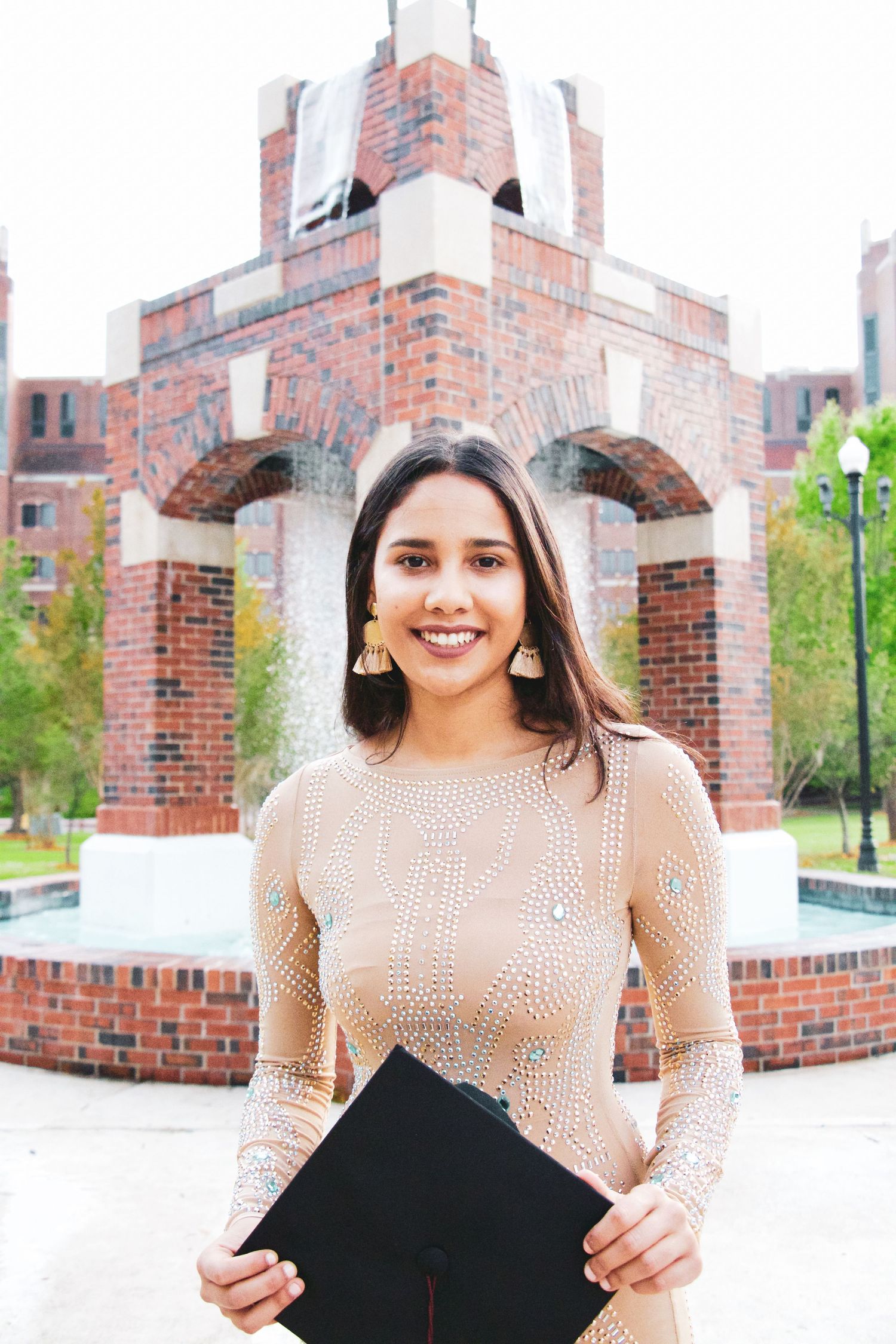 Young woman in beige dress holding graduation cap smiles in front of brick university building.