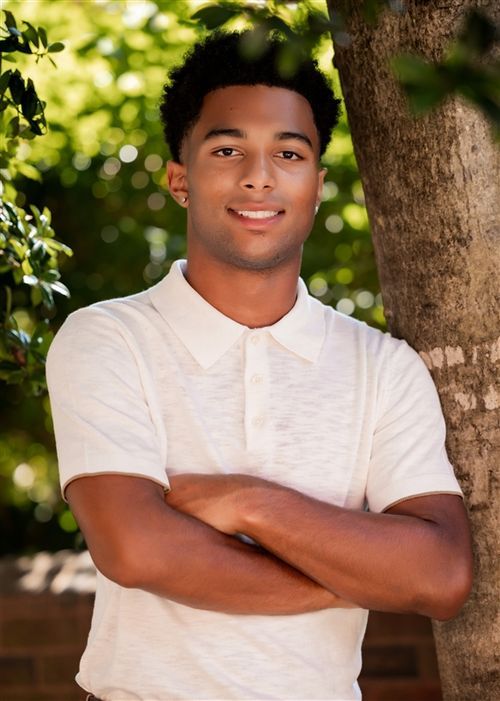 Young man in white polo shirt smiling with arms crossed leaning against tree outdoors.