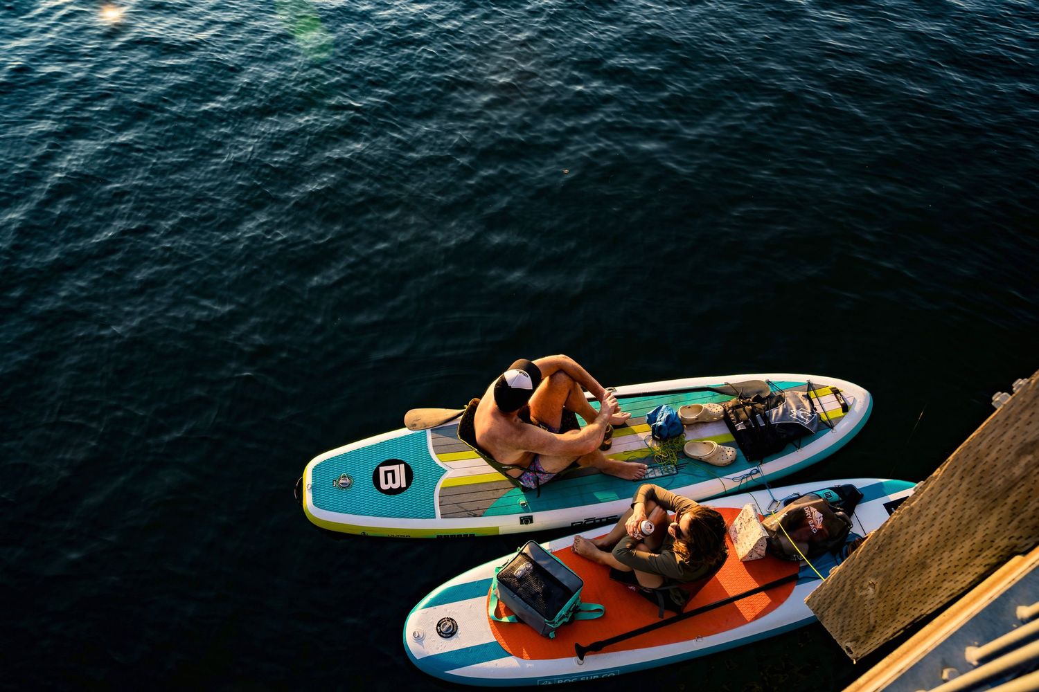 Two people relaxing on colorful paddleboards floating on dark calm water near a dock on a sunny day.