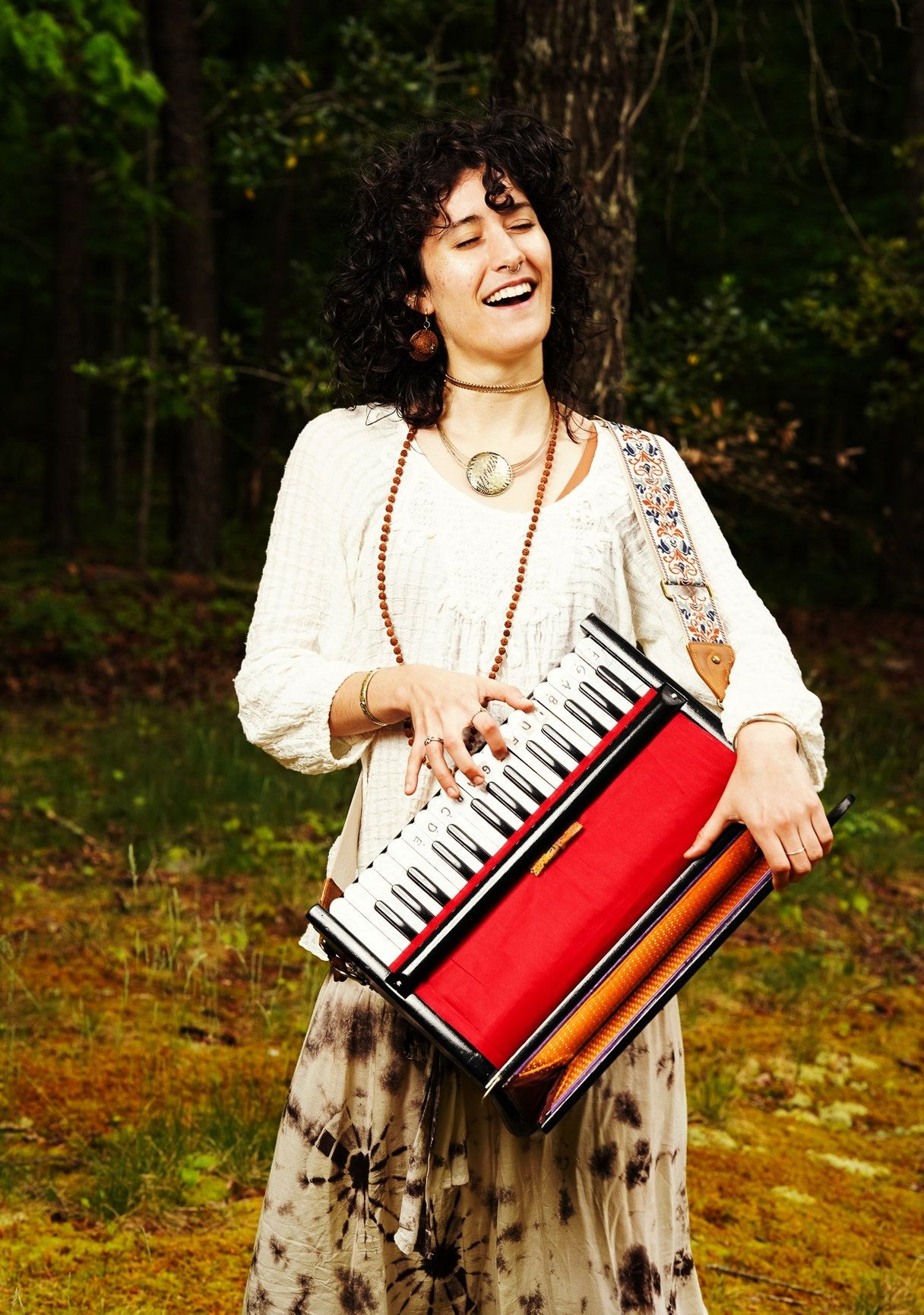 This is a photo of a girl with brown curly hair playing a mini piano, she is wearing a white shirt and has lots of jewelry on.