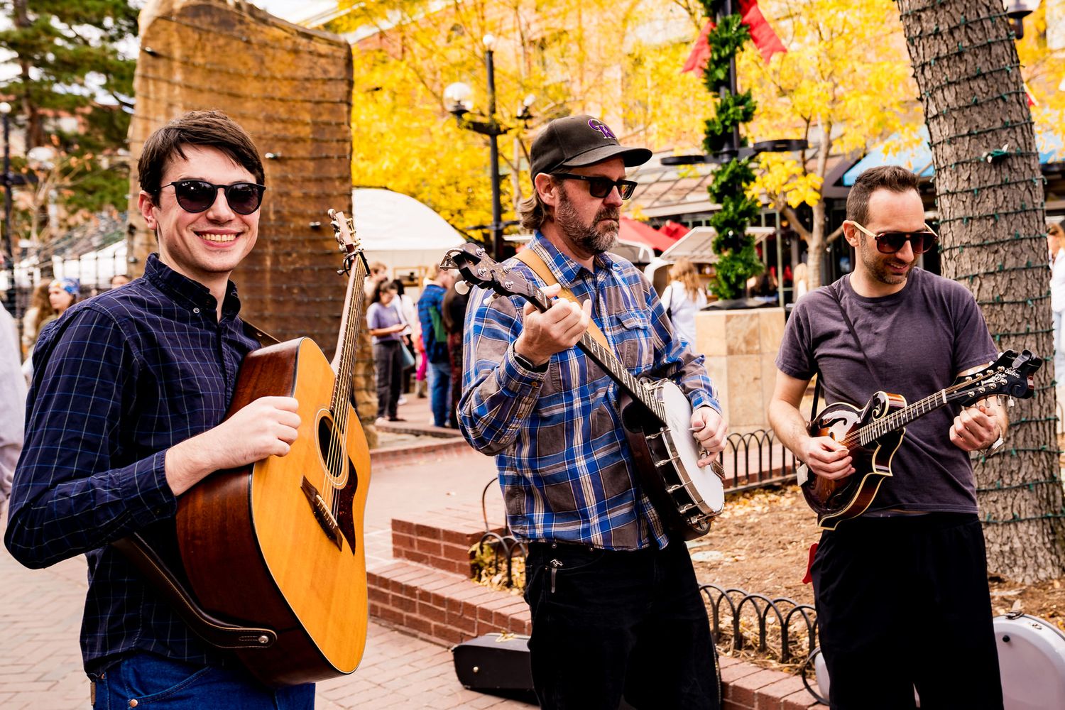 Three musicians playing guitar, banjo, and mandolin outdoors on a city street surrounded by autumn trees.