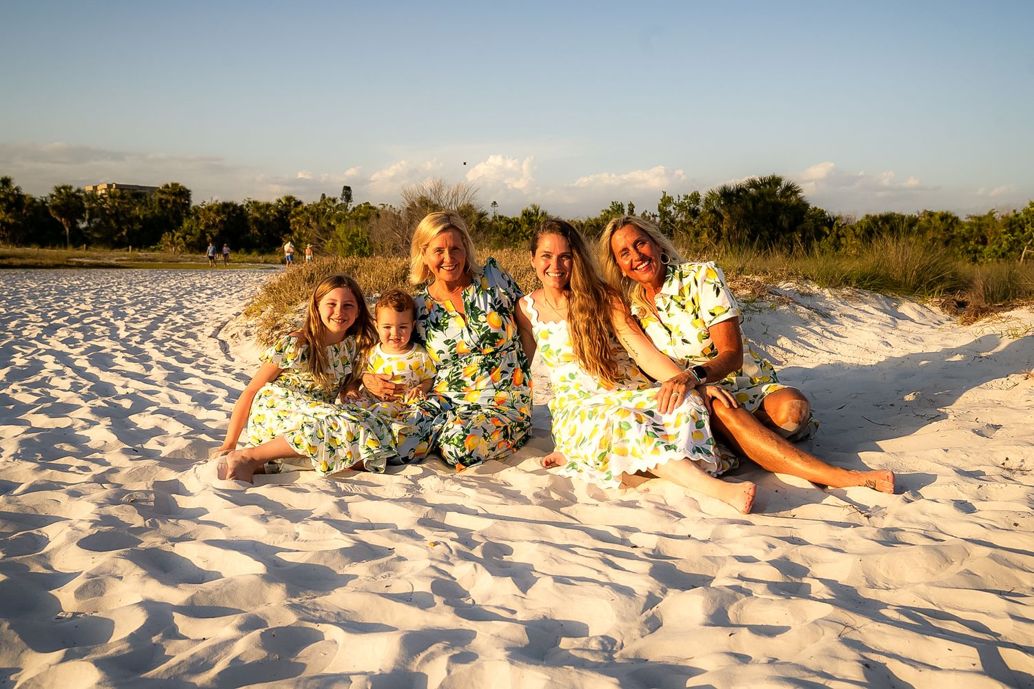 A photo of a family of blonde girls wearing patterned dresses sitting on the sand.
