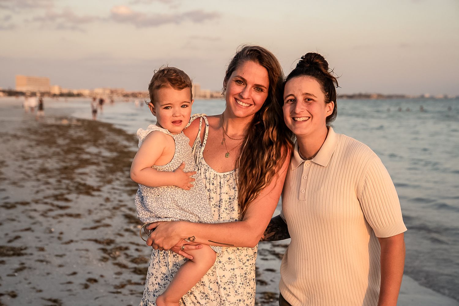 A photo of the two wives and their baby girl on the beach and water behind them with a beautiful sunset.