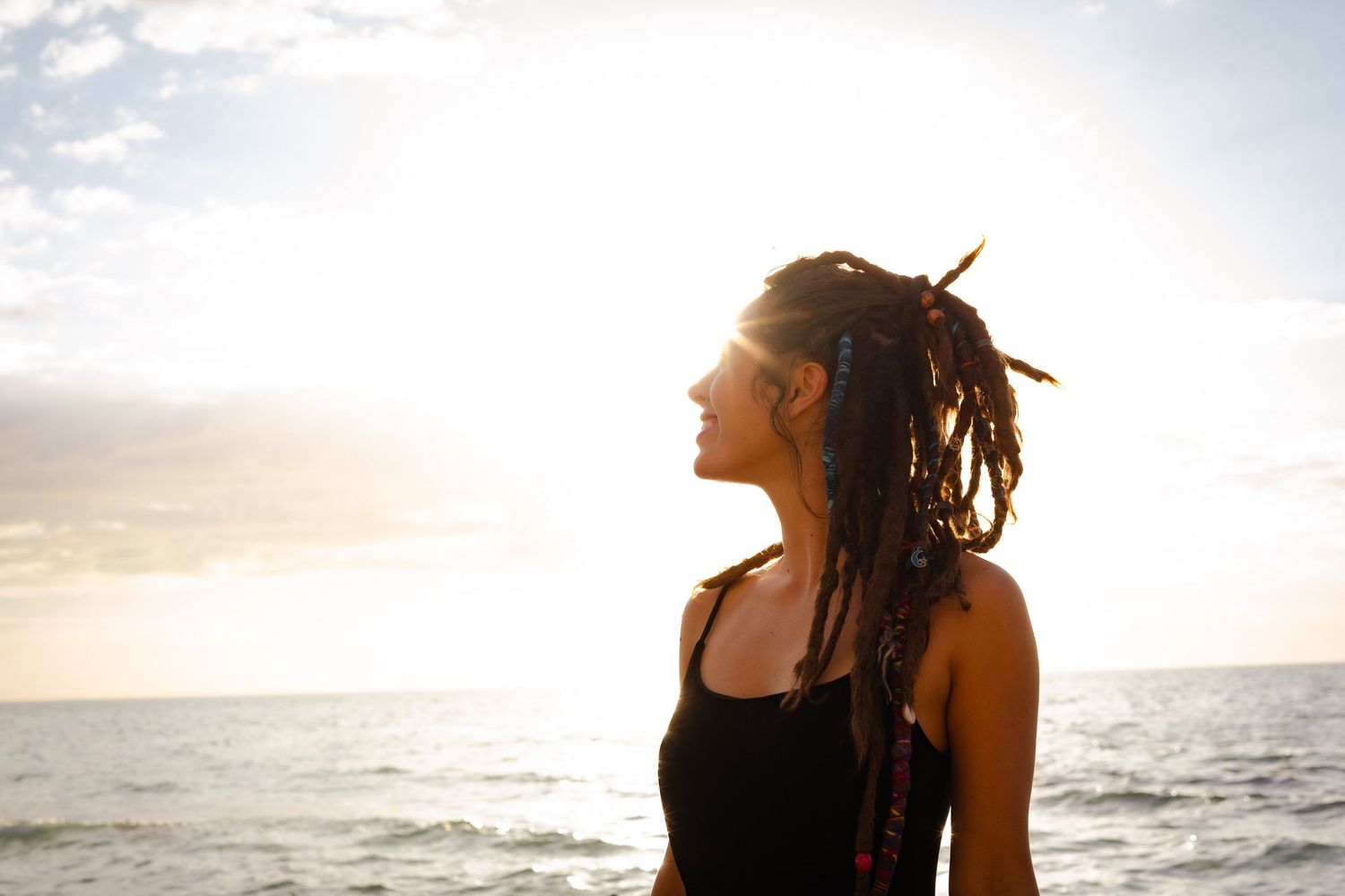 A photo of a woman with dreads and she is looking at the clouds and the sunlight with the water behind her.