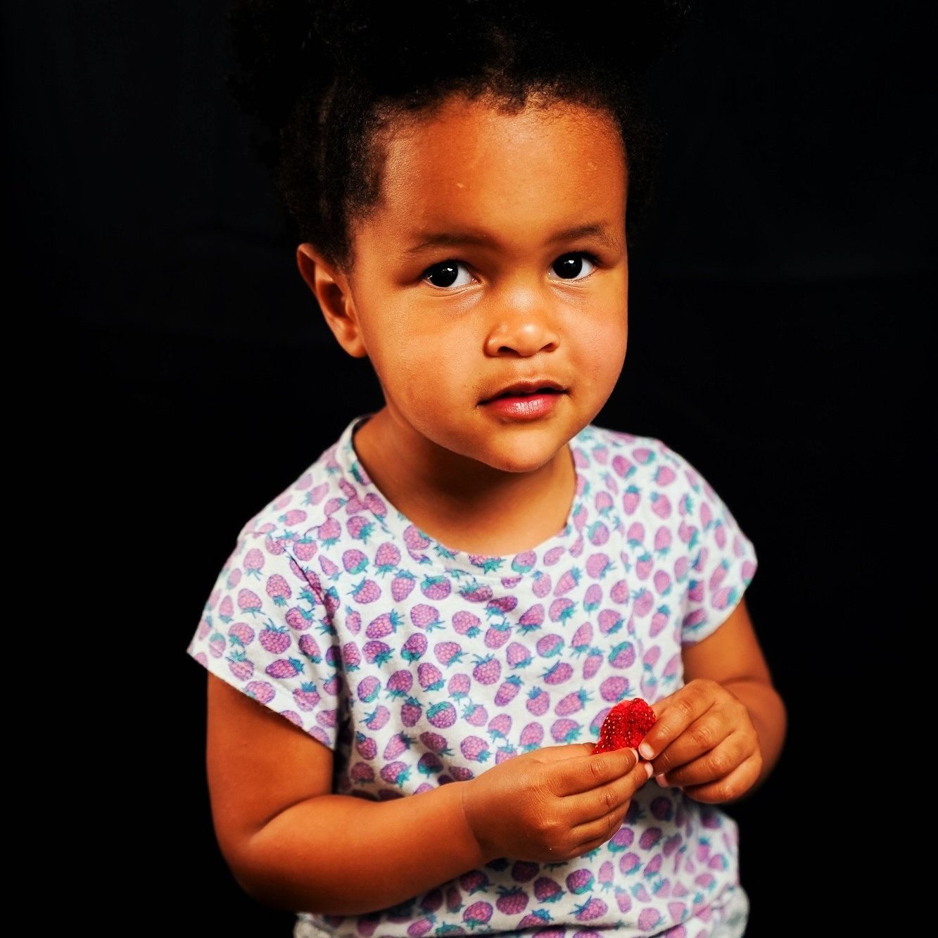 A photo of a young girl looking at the camera and holding a strawberry, she is wearing a strawberry patterned top.
