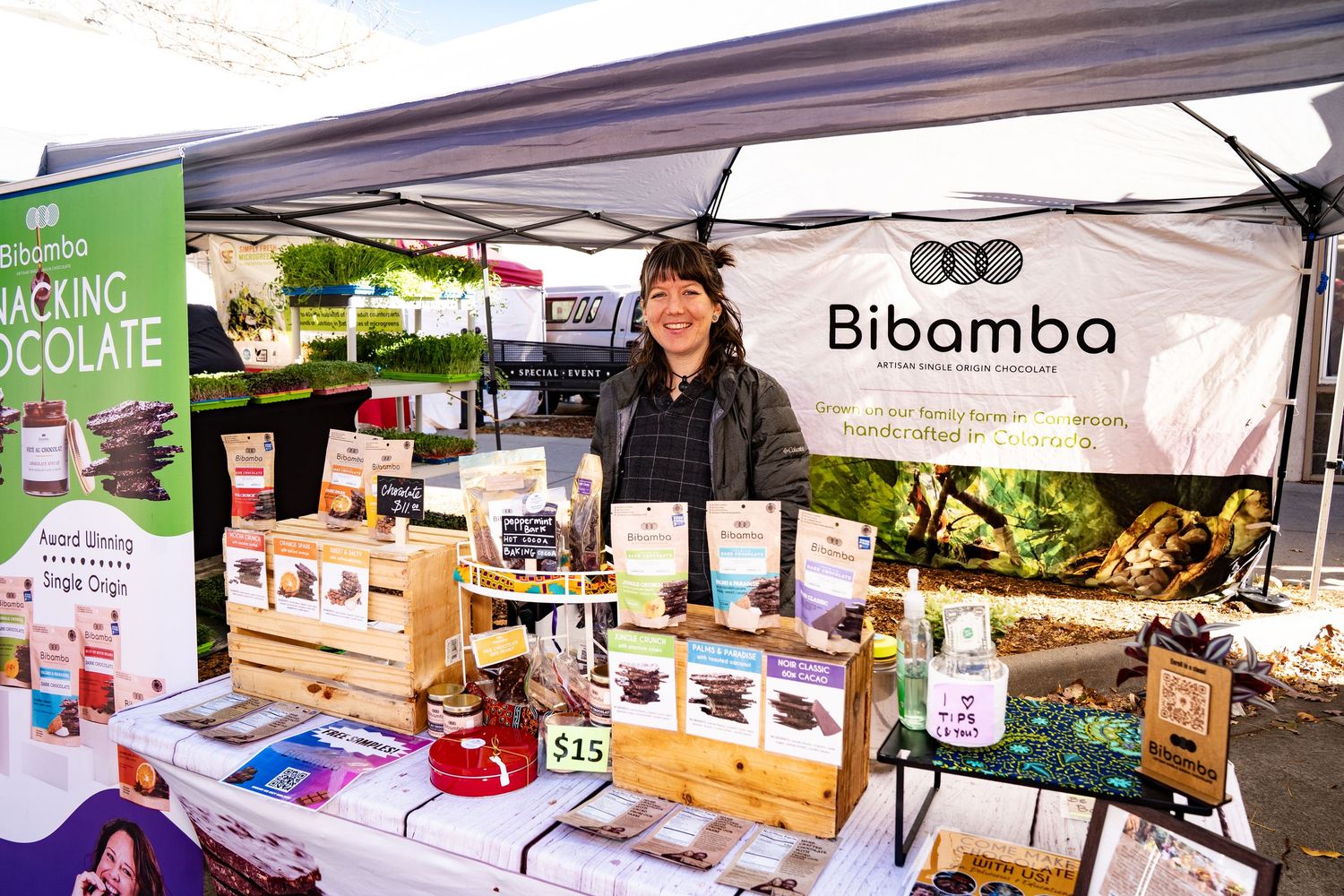 Bibamba Artisan Single Origin Chocolate booth at Boulder Farmers Market with a smiling woman offering chocolate samples, showcasing locally